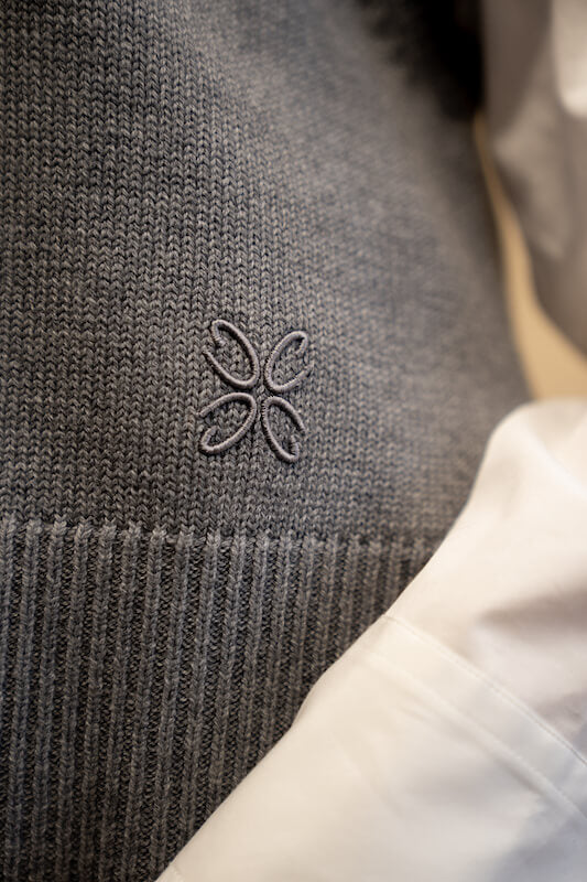 A close-up of gray knitwear from CYME, featuring an embroidered logo. The image also includes a glimpse of a white garment next to the knitwear.