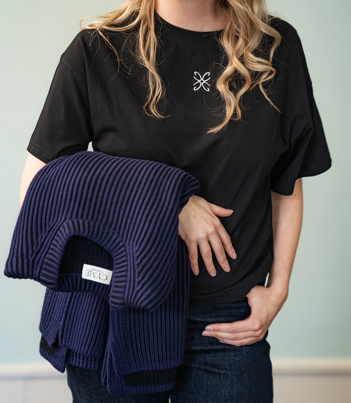 A person wearing a black CYME t-shirt with an embroidered logo, holding a folded navy ribbed sweater with the CYME label visible. The person has long, wavy blonde hair and is dressed in dark jeans.