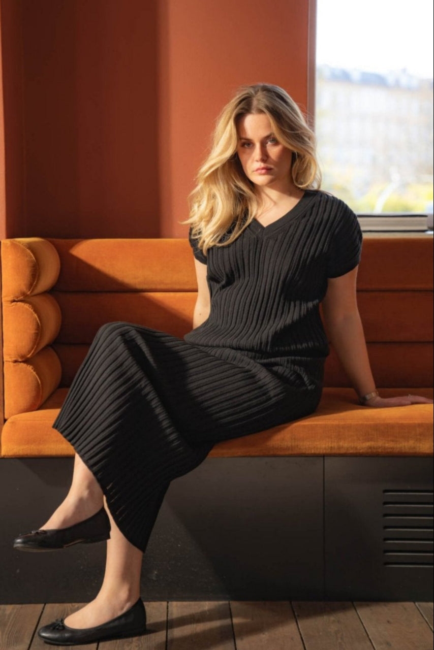 Woman sitting on an orange couch wearing a black ribbed dress by Cyme Copenhagen. She has blonde hair and black ballet flats, with an orange and sunlit background. The scene highlights the elegance and comfort.