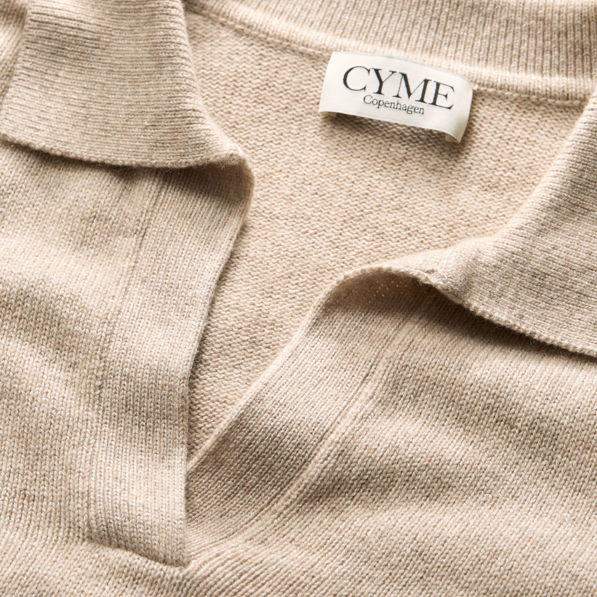 A close-up of the collar and neckline of a beige polo-style knit jumper, featuring a "CYME Copenhagen" label.