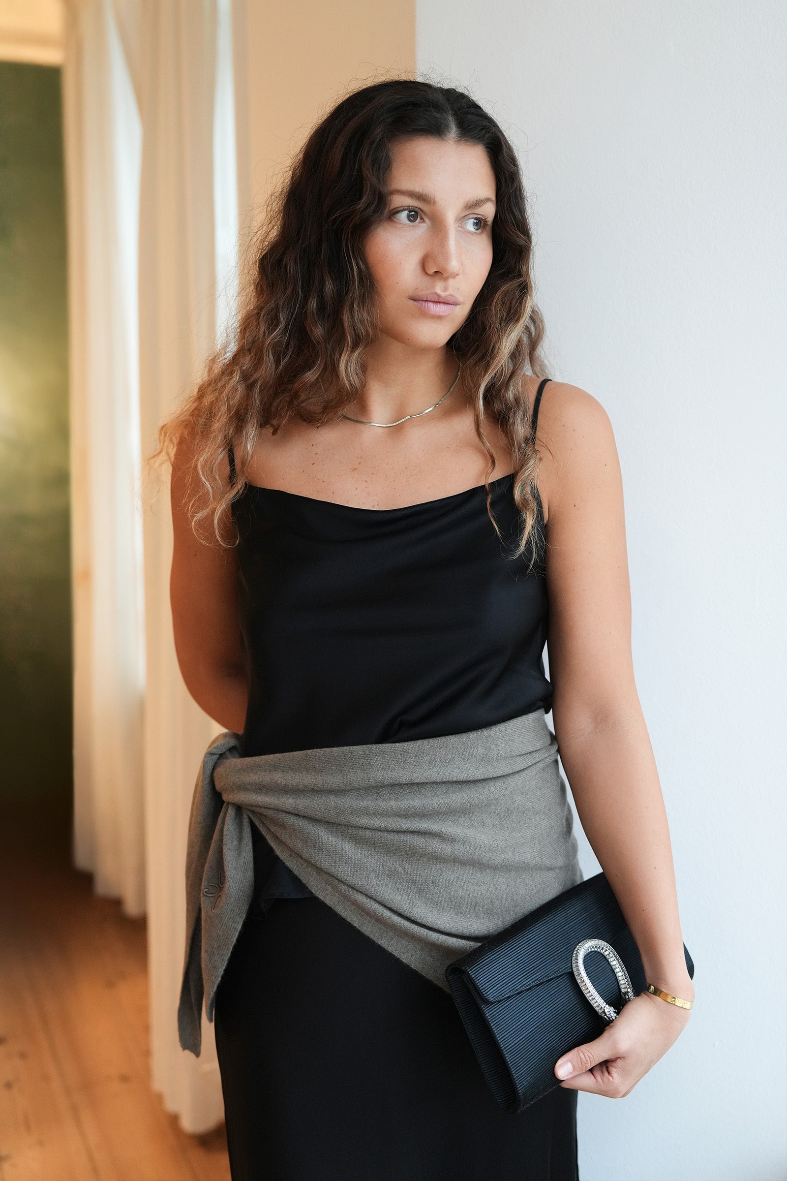 Woman wearing a black top and gray wrap holding a black clutch indoors.