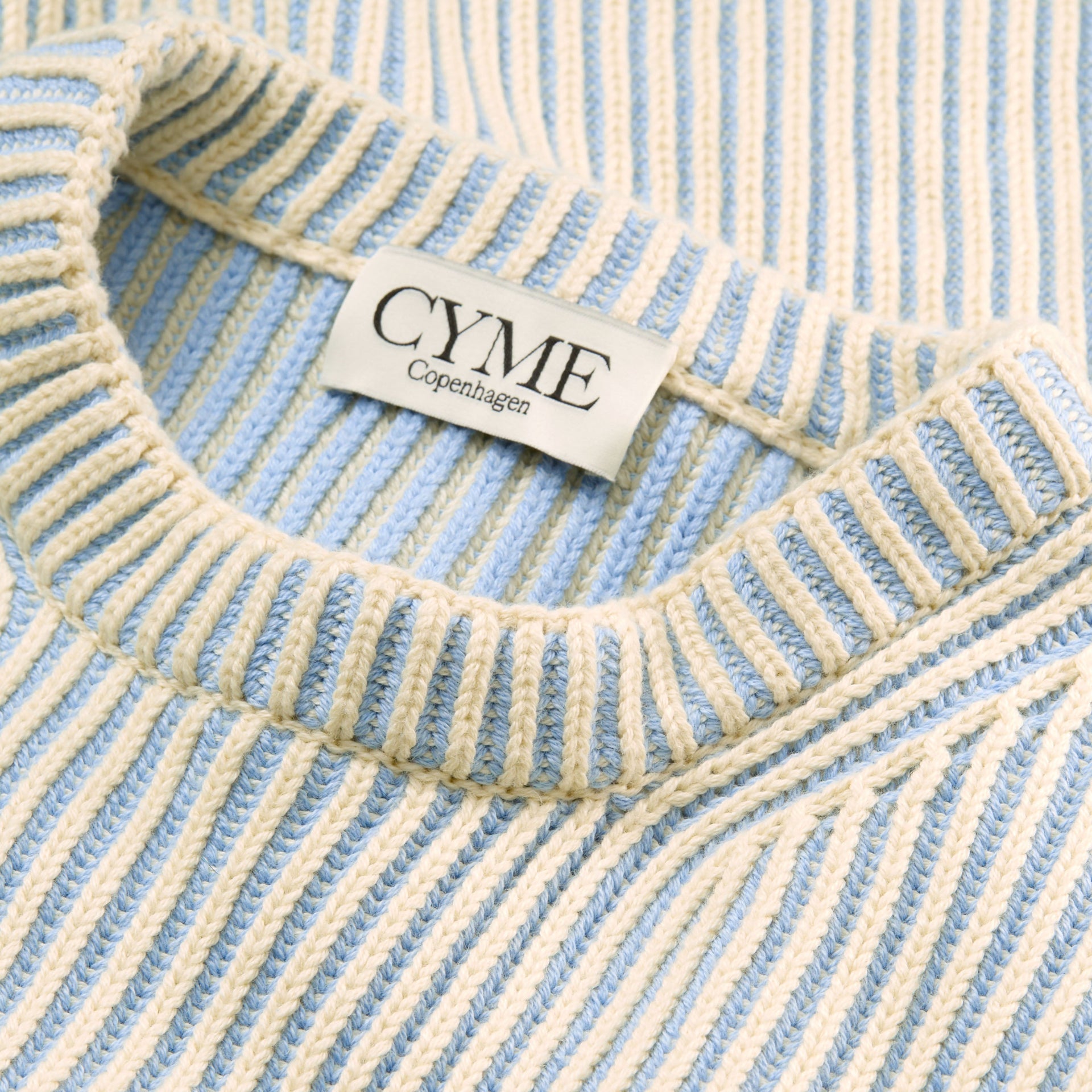 A close-up of the neckline and ribbed texture of a cream and light blue sweater, with a visible "CYME Copenhagen" label.