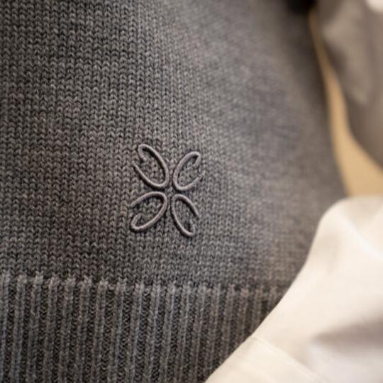 A close-up of gray knitwear from CYME, featuring an embroidered logo. The image also includes a glimpse of a white garment next to the knitwear.