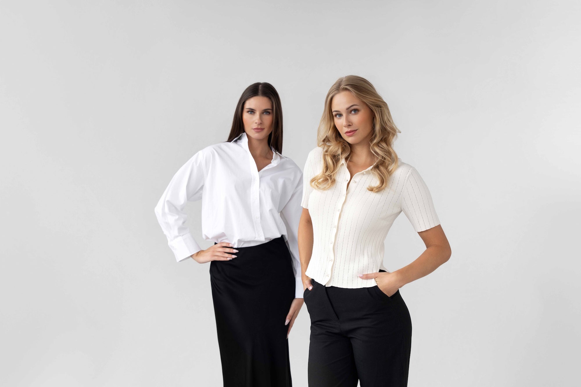Two women wearing white shirts and black pants standing in a studio setting.