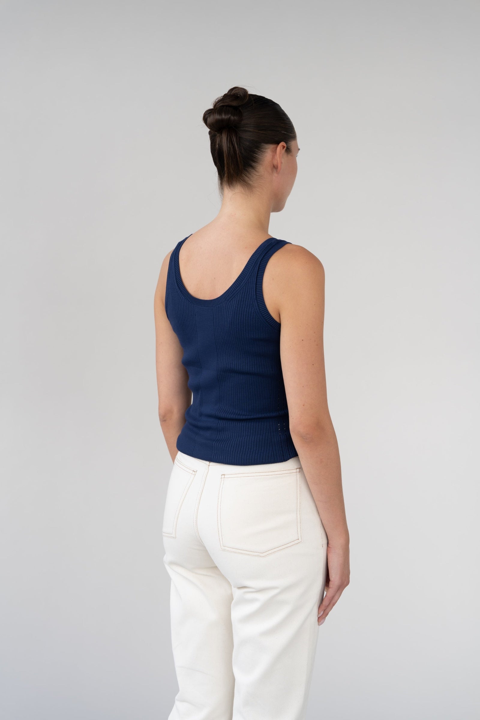 Woman wearing a navy tank top and white pants against a gray background