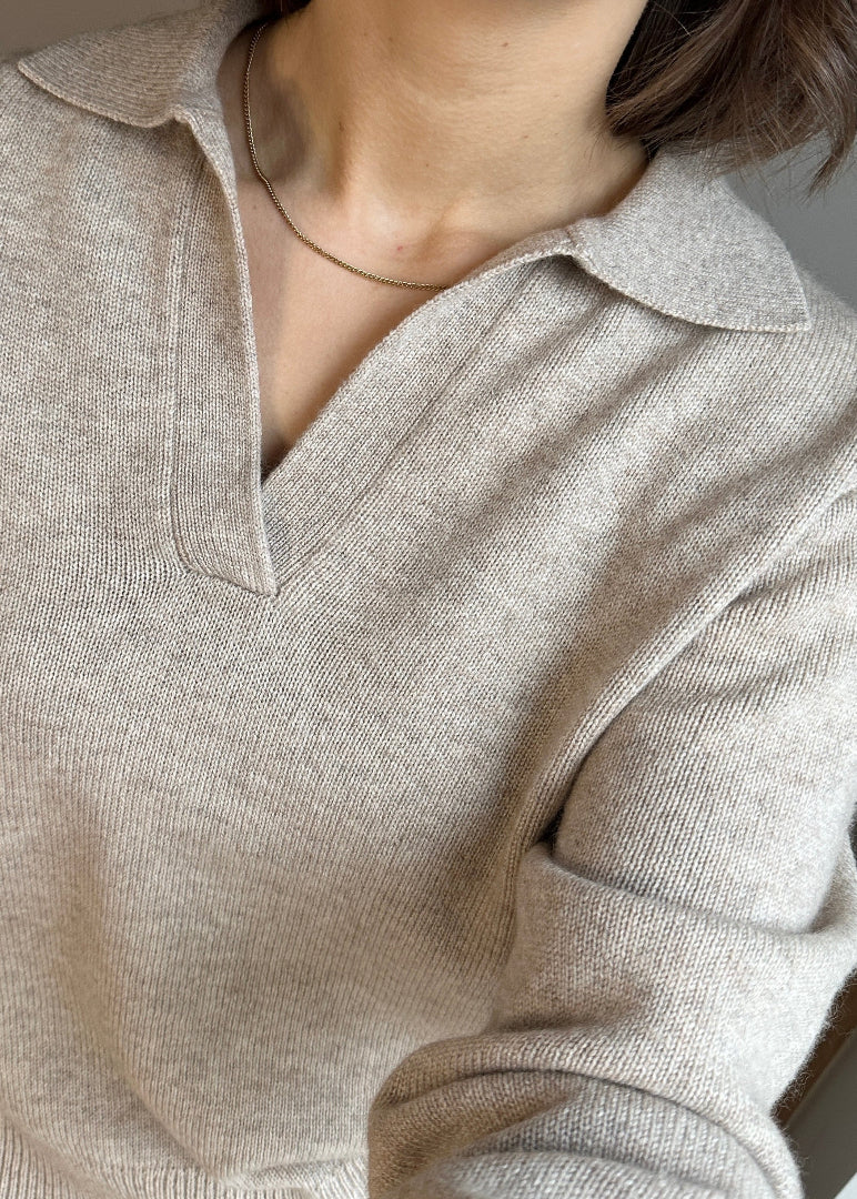 A close-up of a beige polo-style knit jumper with a V-neck and collar, worn by a person with a gold necklace.