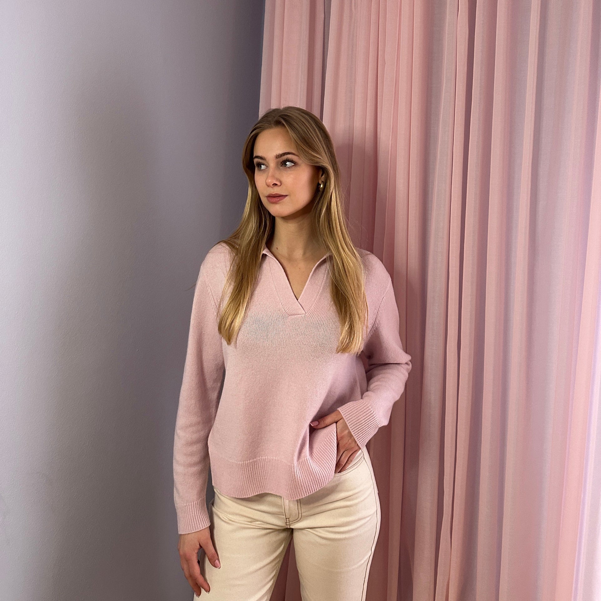 Woman wearing a light pink polo-style knit jumper, standing against pink curtains.