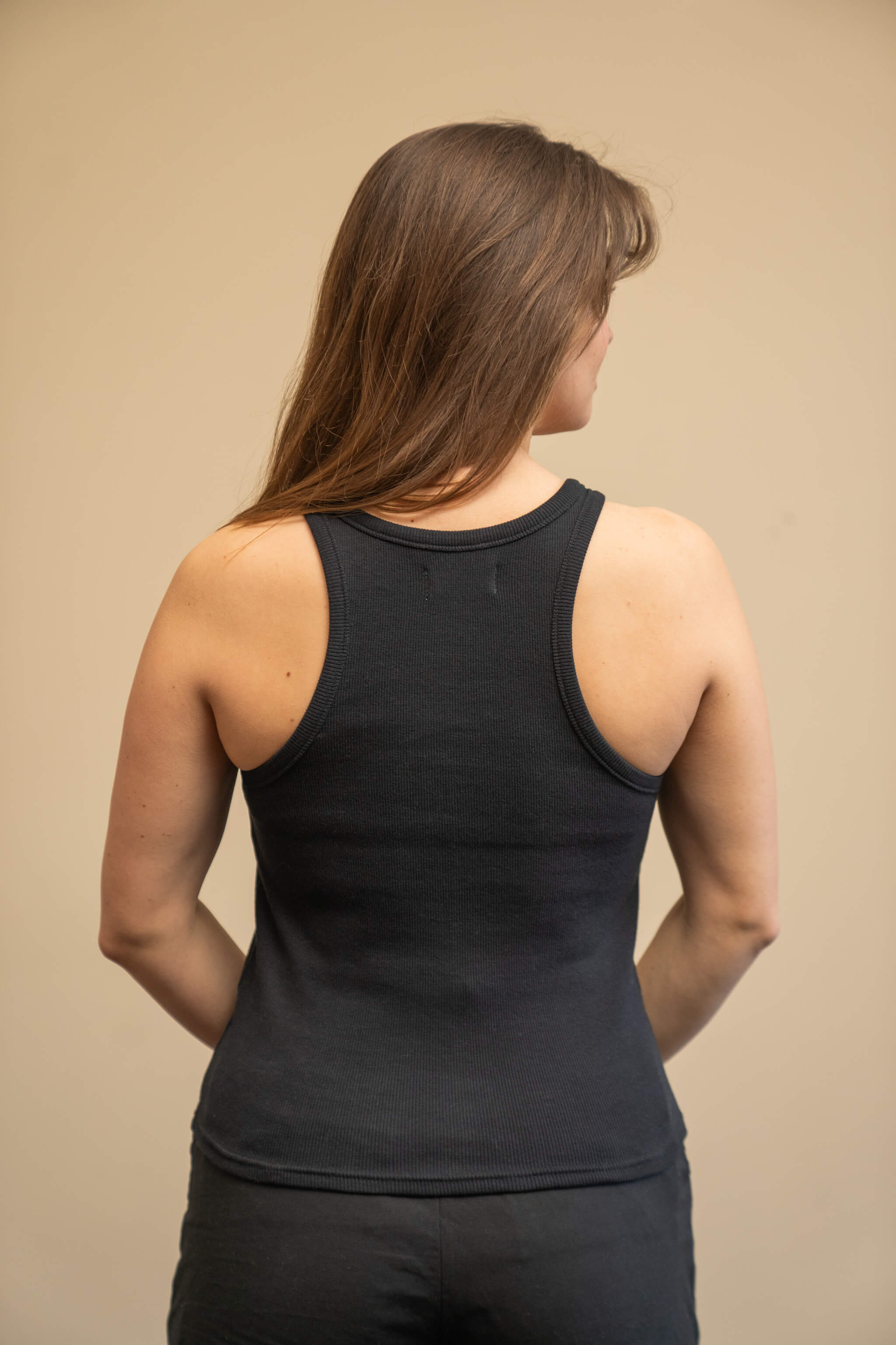 Rear view of a woman wearing a black tank top from Cyme Copenhagen's spring/summer collection. The tank top features a simple and sleek design with racerback styling. The model stands against a neutral background, highlighting the clean lines and minimalist appeal of the garment.