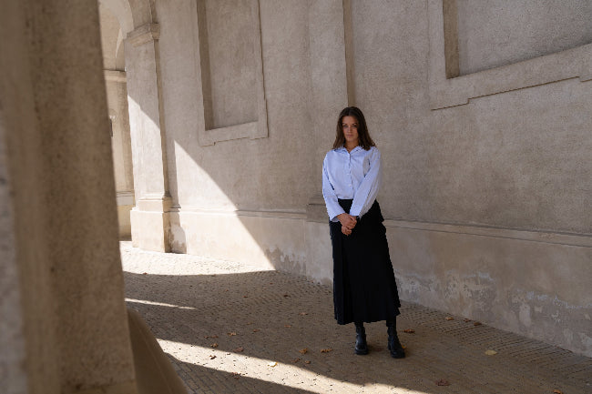 Woman in a white shirt and black skirt standing in a sunlit hallway with stone walls.