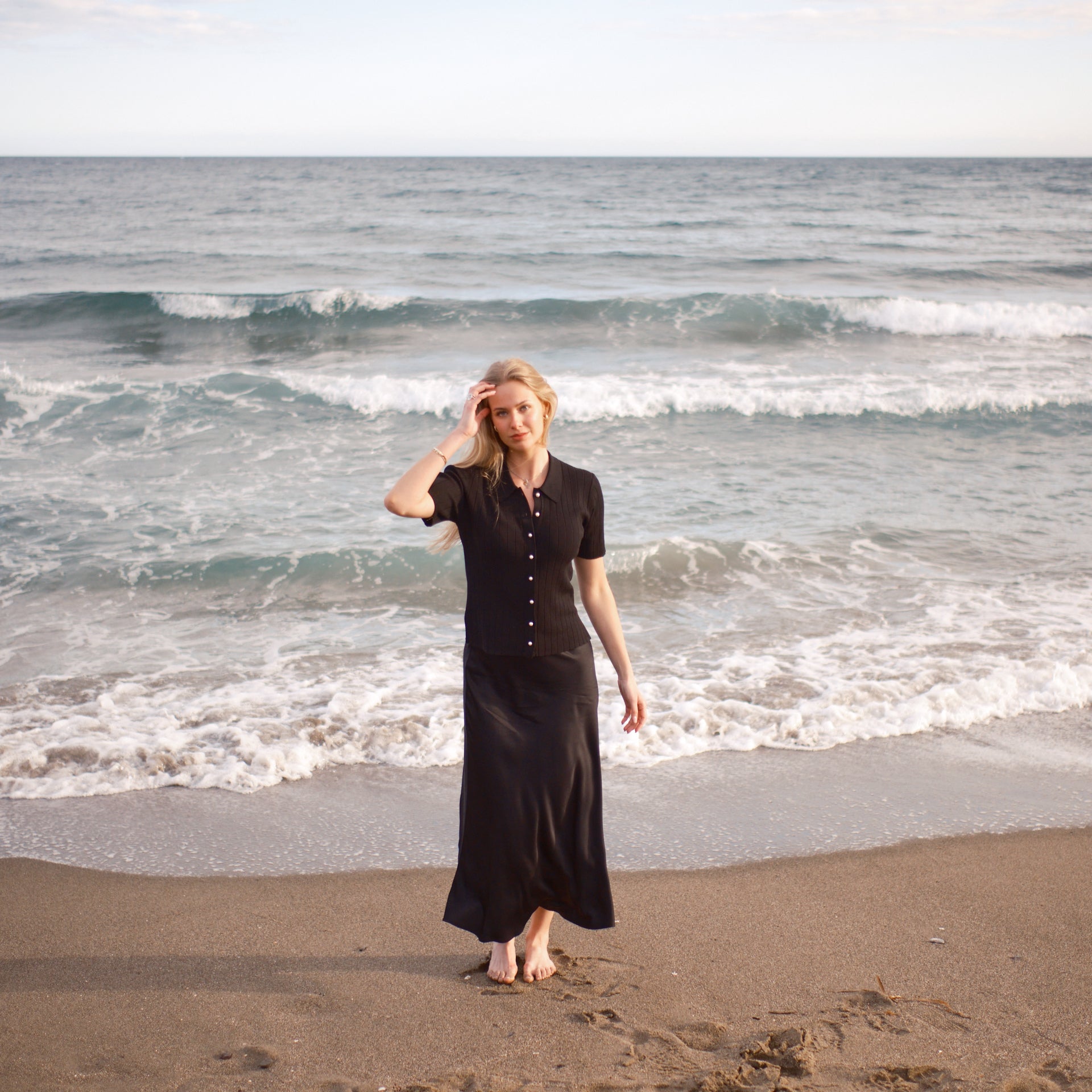 Model iført sort ribstrikket cardigan med perleknapper og lang silkeskørt fra CYME Copenhagen, gående barfodet langs stranden med bølger i baggrunden. Et elegant og roligt slow fashion-look i naturlige omgivelser.