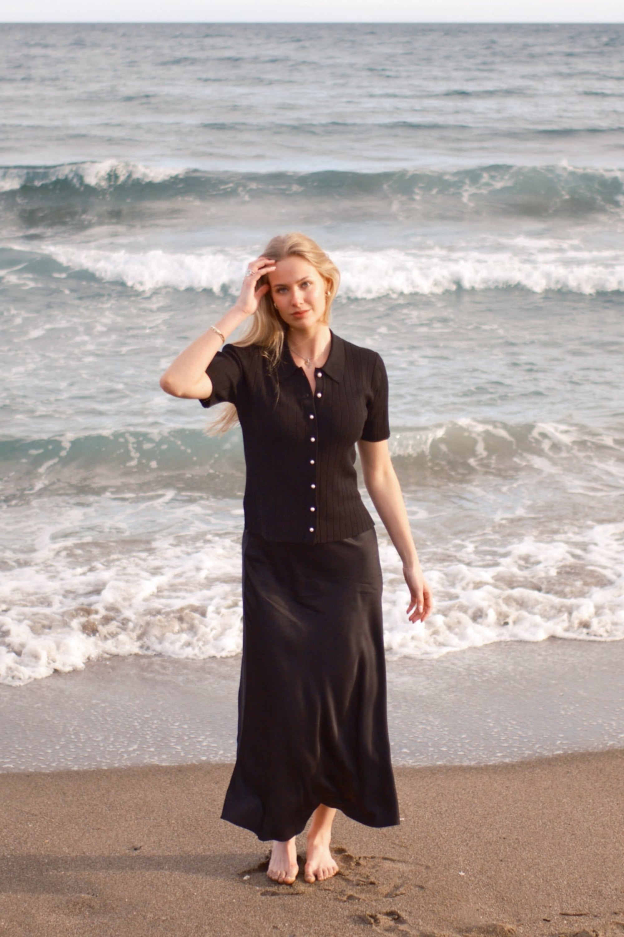 Model iført sort ribstrikket cardigan med perleknapper og lang silkeskørt fra CYME Copenhagen, gående barfodet langs stranden med bølger i baggrunden. Et elegant og roligt slow fashion-look i naturlige omgivelser.