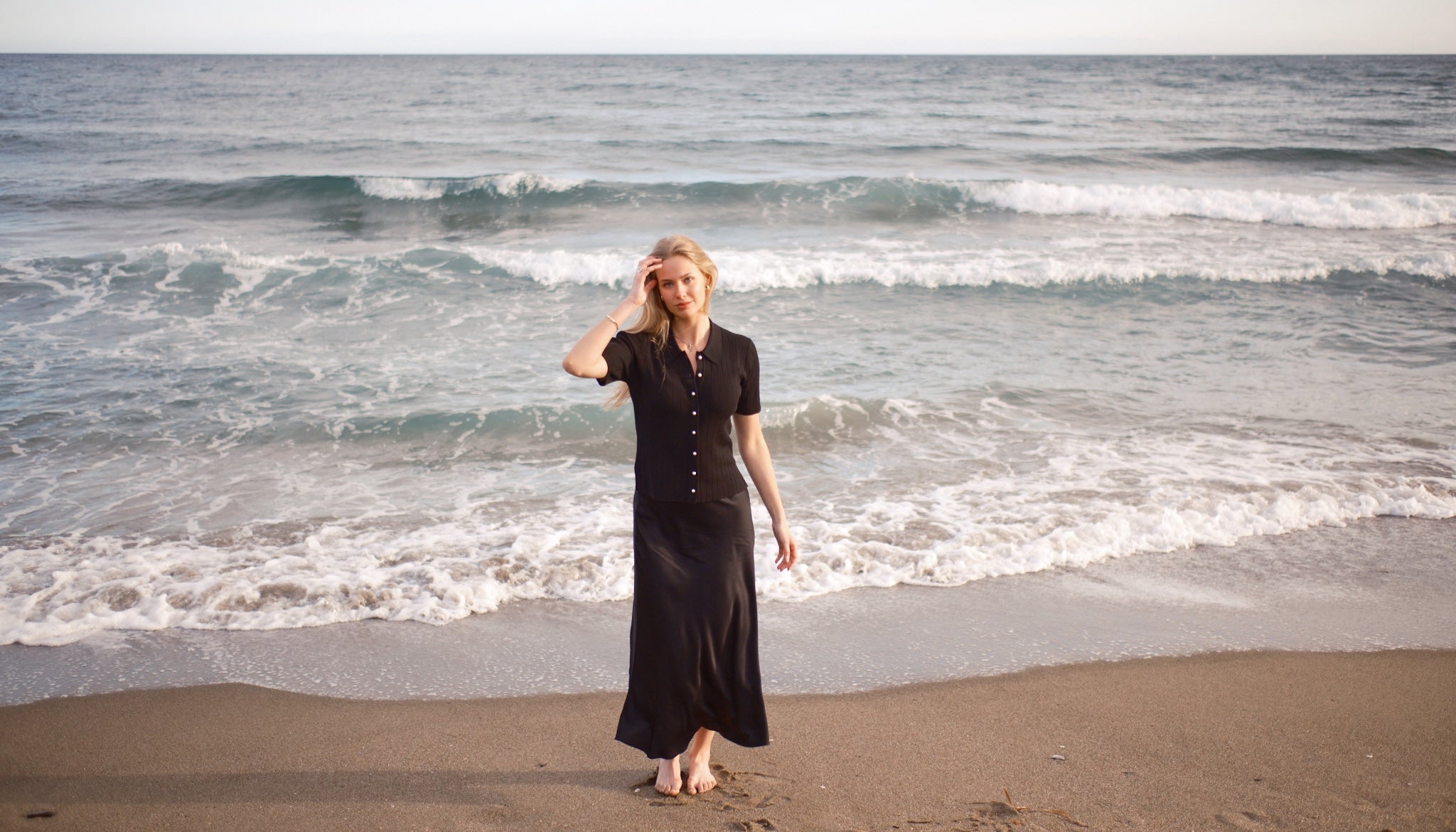 Woman in black rib-knit pearl-button top and satin maxi skirt, barefoot on a sandy beach with gentle waves. Cyme Copenhagen landing hero.