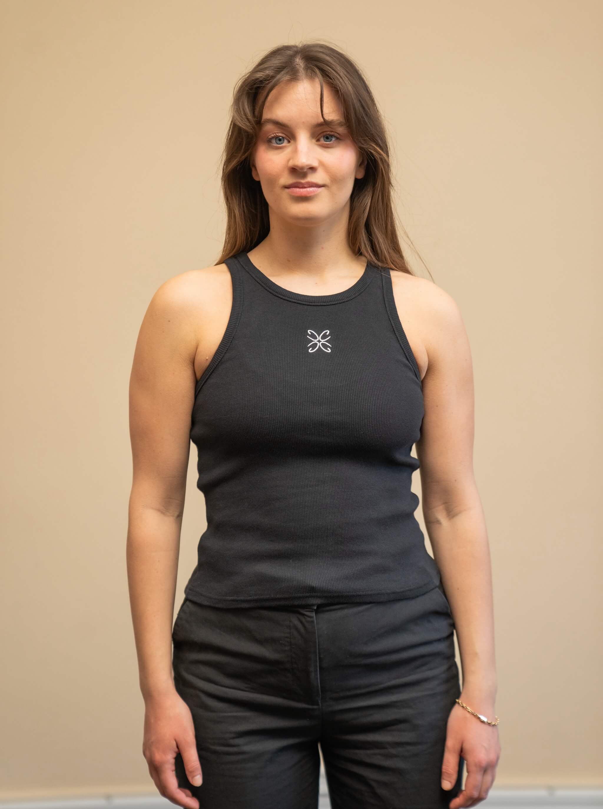 Woman wearing a black tank top from Cyme Copenhagen's spring/summer collection. The tank top features a simple design with a white logo in the center. The model stands against a neutral background, emphasizing the minimalist and stylish appeal of the garment.