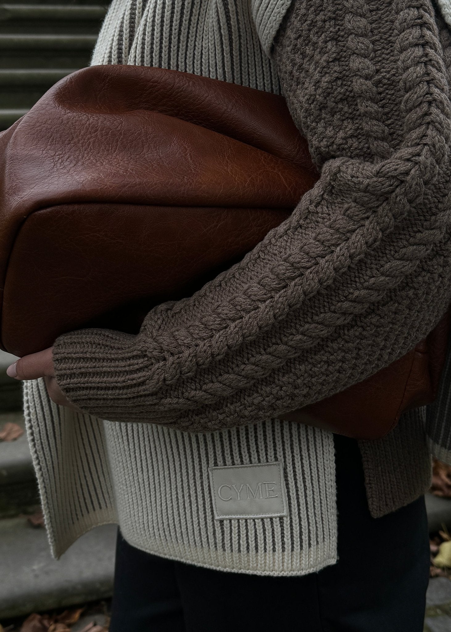 Close-up of a CYME Copenhagen cable knitted cardigan, paired with a brown leather bag, showcasing premium craftsmanship.

