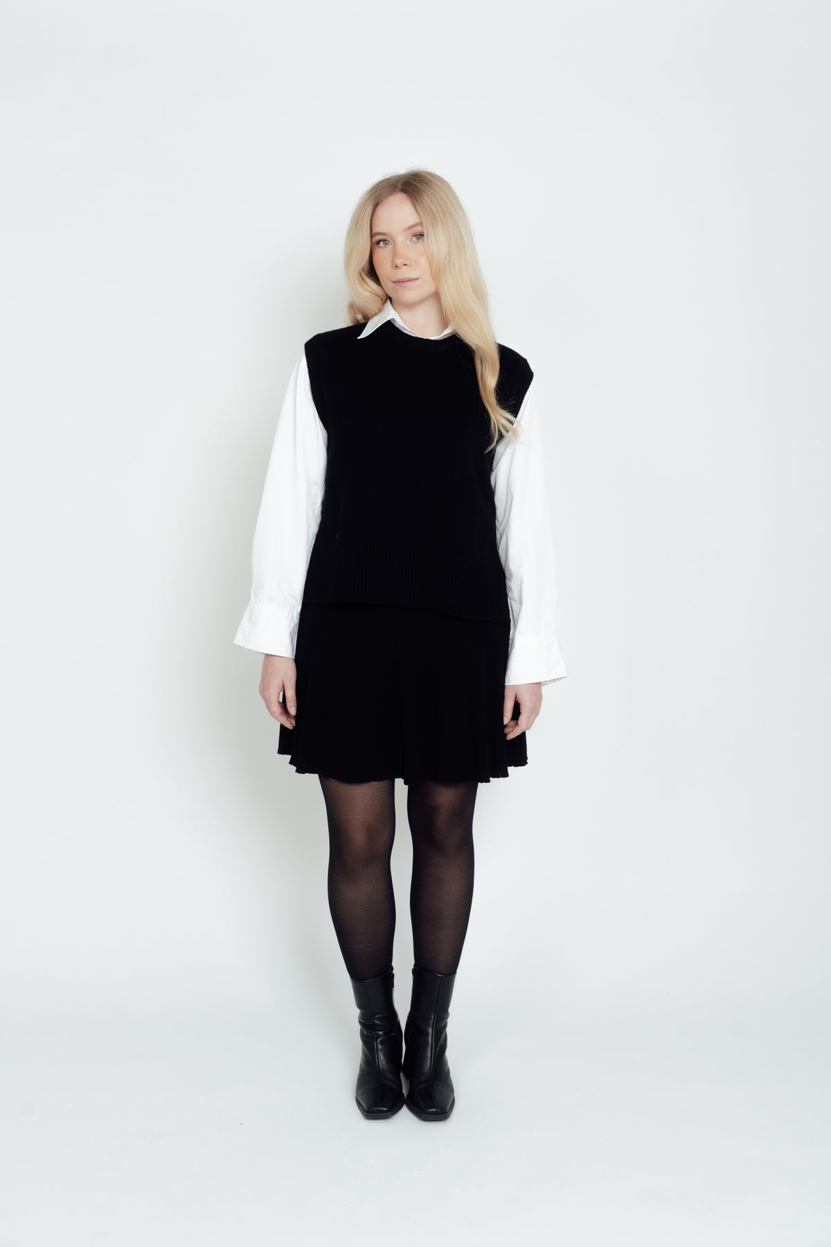 Full-length view of a woman wearing the black CYME Merino Crewneck Vest with a white shirt, black skirt and boots.