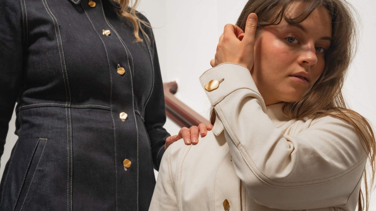 Close-up of two women wearing denim jackets from Cyme Copenhagen's spring/summer 2024 collection. The woman on the left wears a dark denim jacket with gold buttons, while the woman on the right wears a light denim jacket with matching gold buttons. The focus is on the detailed stitching and button design, highlighting the brand's attention to craftsmanship and style.