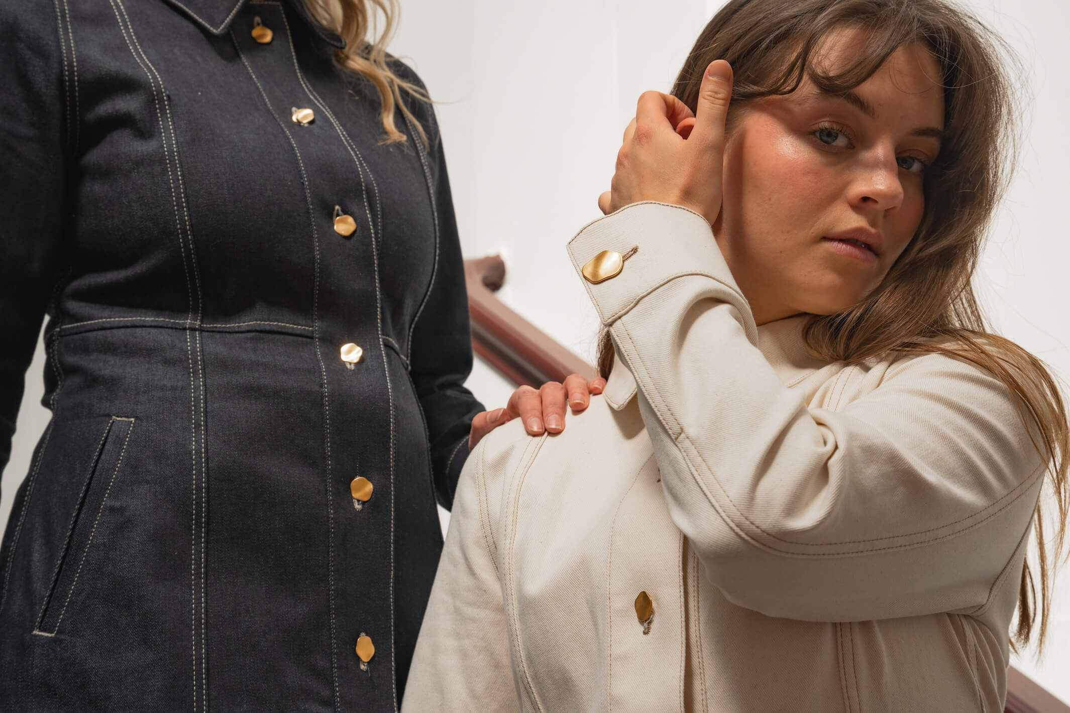 Close-up of two women wearing denim jackets from Cyme Copenhagen's spring/summer 2024 collection. The woman on the left wears a dark denim jacket with gold buttons, while the woman on the right wears a light denim jacket with matching gold buttons. The focus is on the detailed stitching and button design, highlighting the brand's attention to craftsmanship and style.