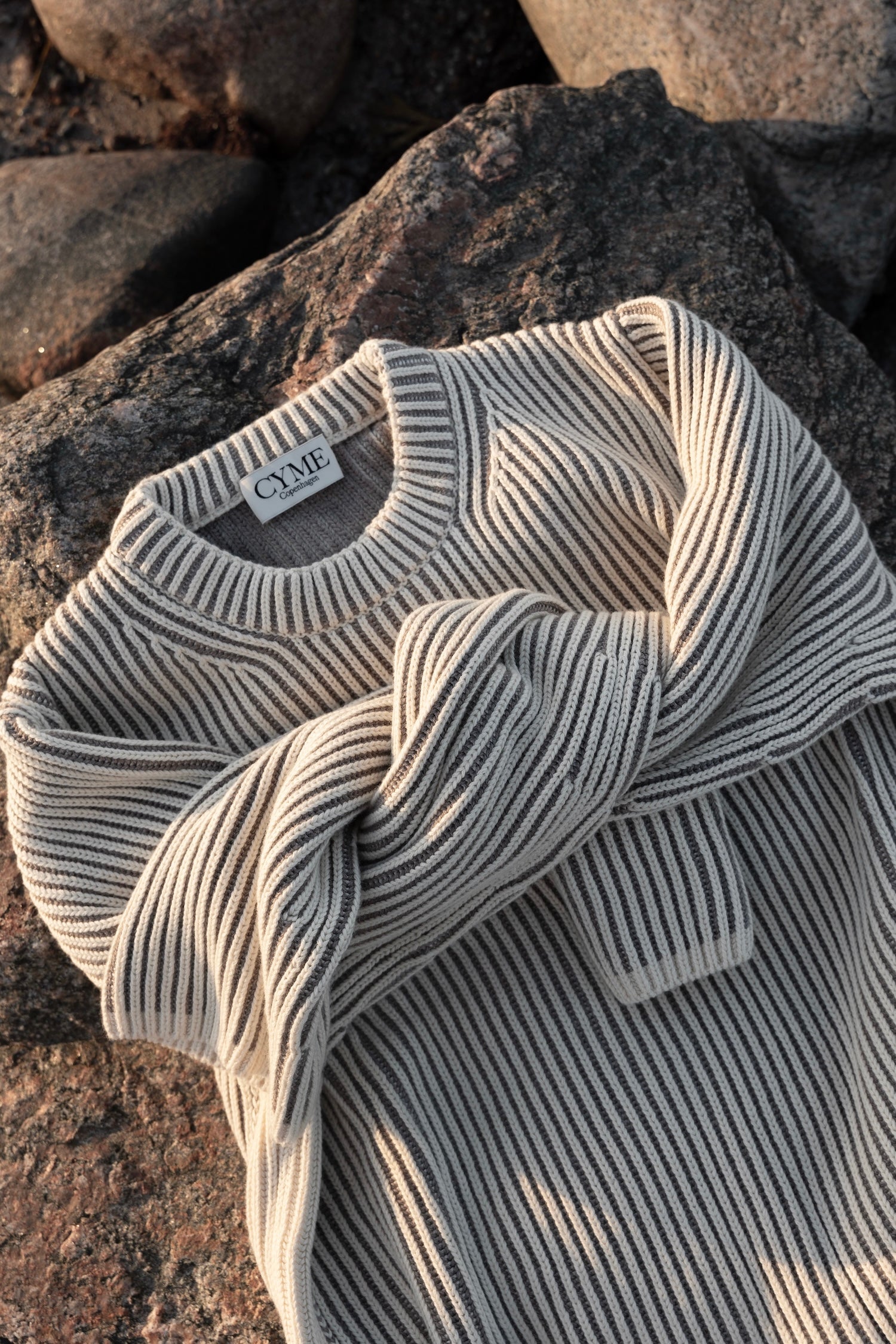 Cream CYME cotton-cashmere jumper with contrast ribbing laid across sunlit coastal rocks.