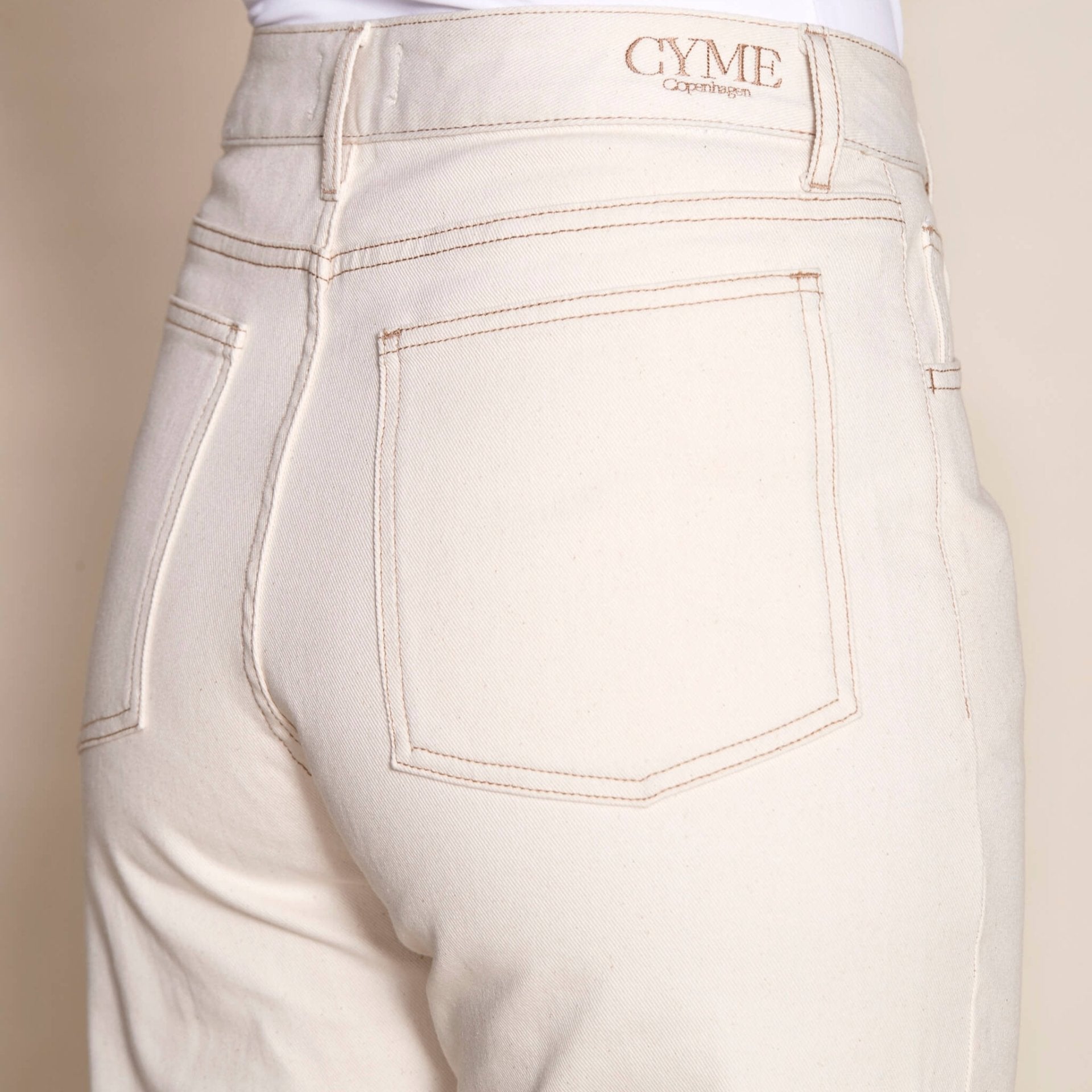 Detail of Cyme Copenhagen's cream denim pants, focusing on the back pocket with branded tag, emphasizing the brand's quality craftsmanship and ethical fashion standards in Denmark's clothing shops.