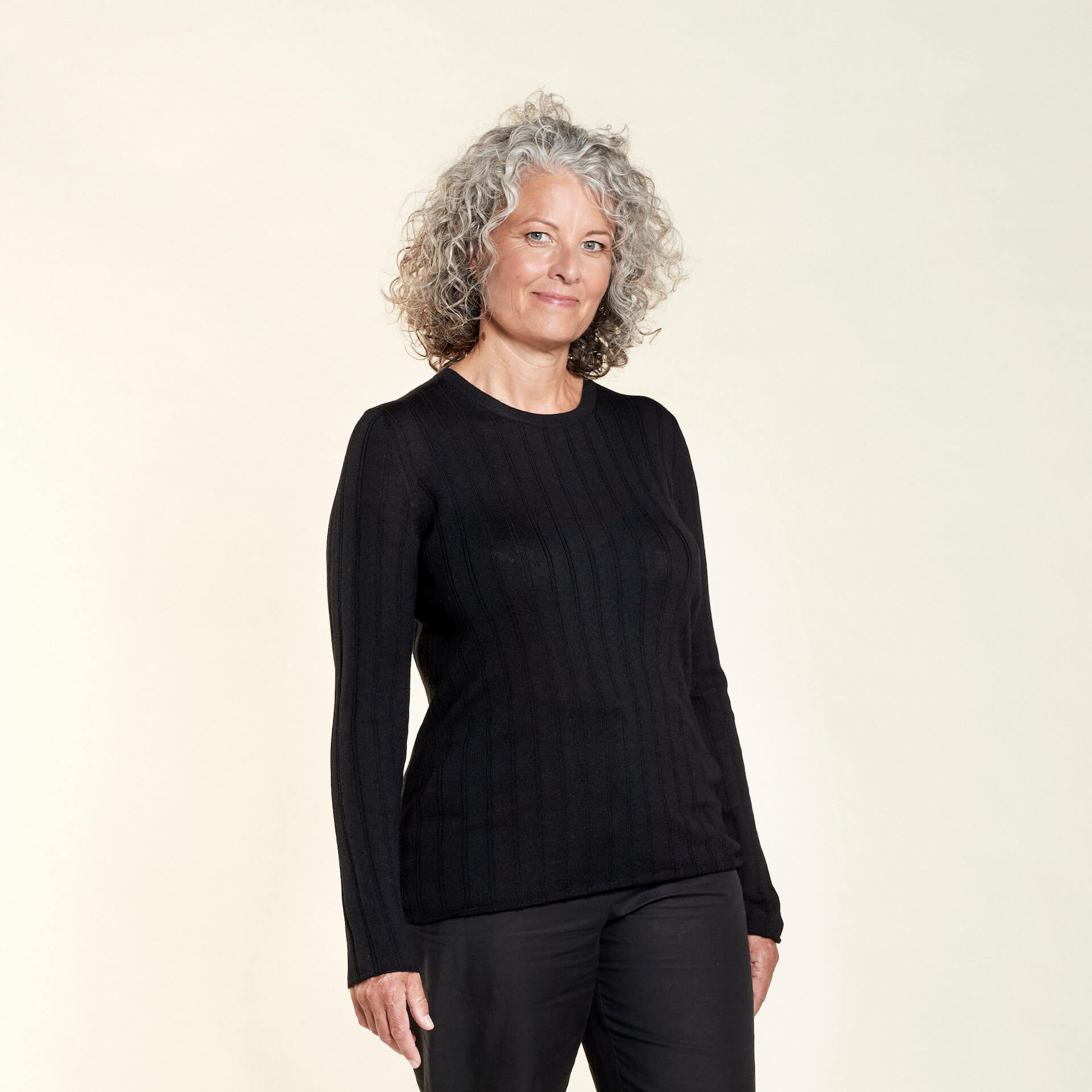 A woman with curly gray hair models a black ribbed long-sleeve top paired with black trousers from CYME Copenhagen. The outfit highlights the brand's focus on timeless, sophisticated, and high-quality fashion.