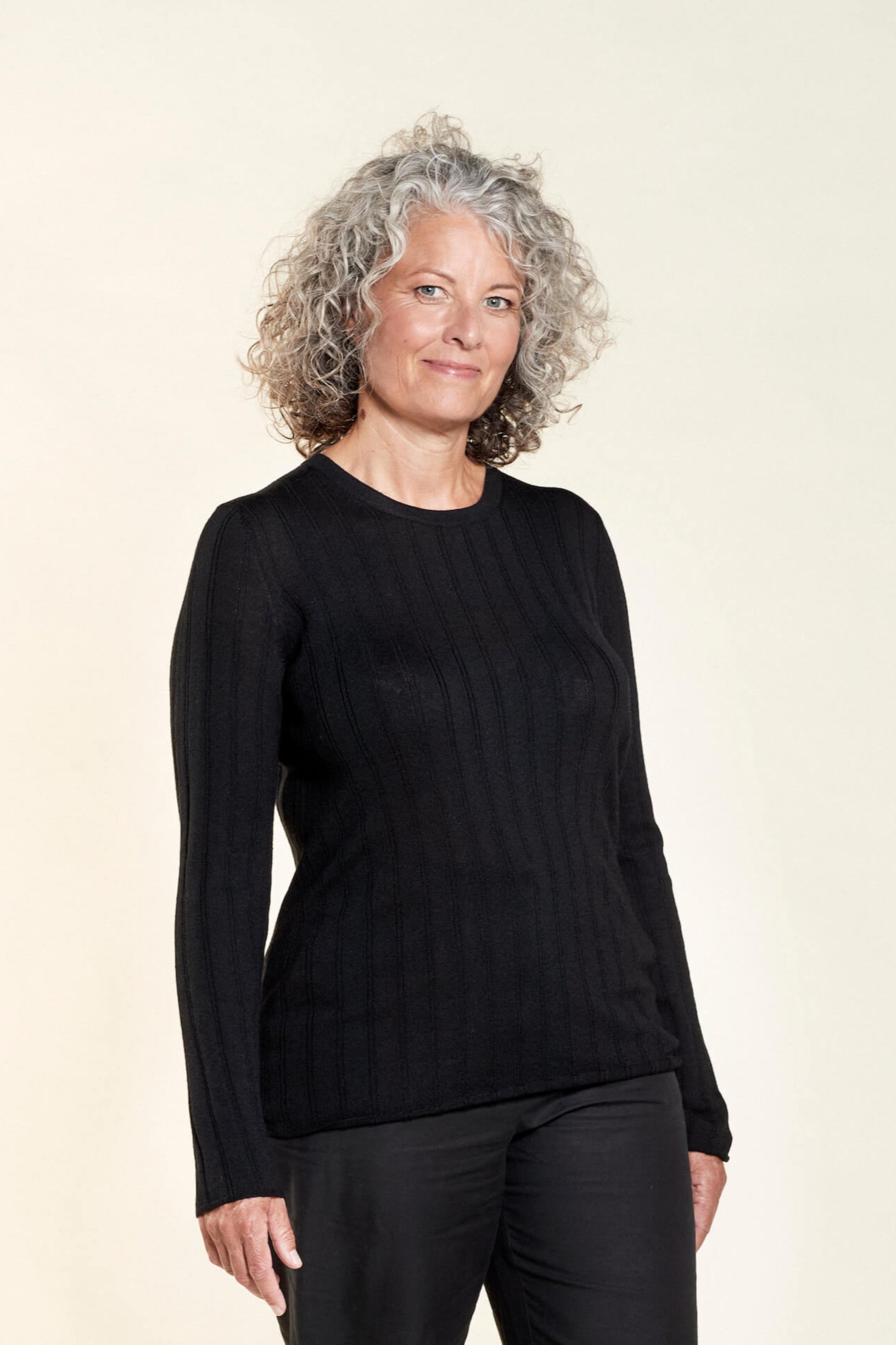 A woman with curly gray hair models a black ribbed long-sleeve top paired with black trousers from CYME Copenhagen. The outfit highlights the brand's focus on timeless, sophisticated, and high-quality fashion.