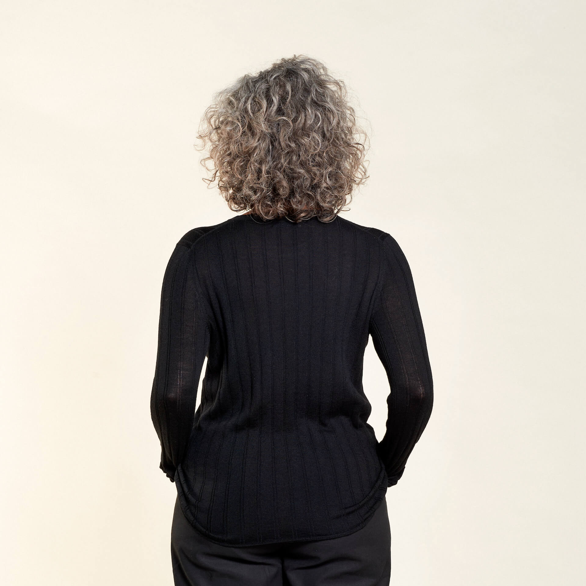 Rear view of a woman with curly gray hair modeling a black ribbed long-sleeve top paired with black trousers from CYME Copenhagen. The image highlights the top's fitted design and the clean lines of the outfit, emphasizing the brand's focus on timeless and versatile fashion.