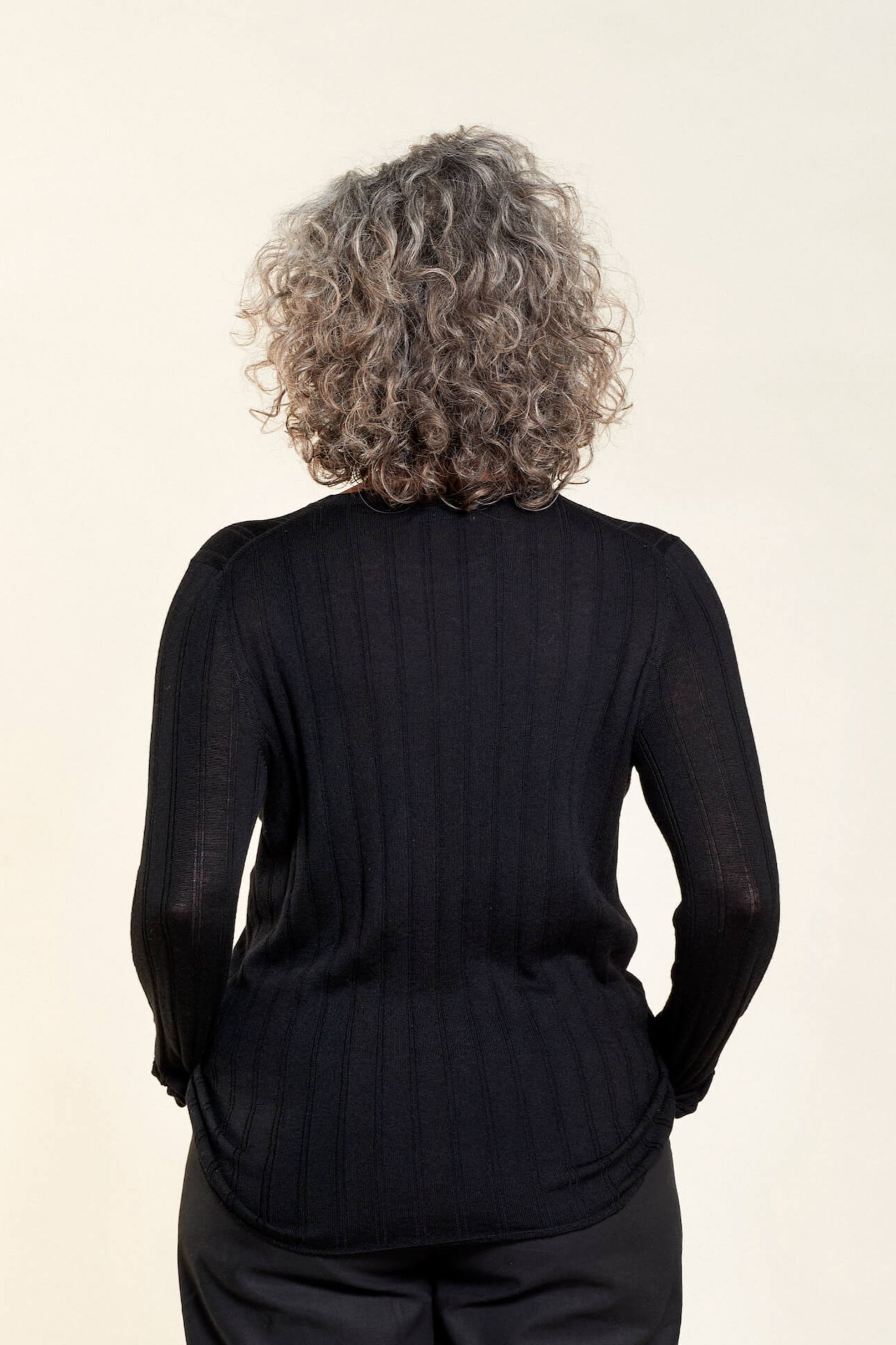 Rear view of a woman with curly gray hair modeling a black ribbed long-sleeve top paired with black trousers from CYME Copenhagen. The image highlights the top's fitted design and the clean lines of the outfit, emphasizing the brand's focus on timeless and versatile fashion.