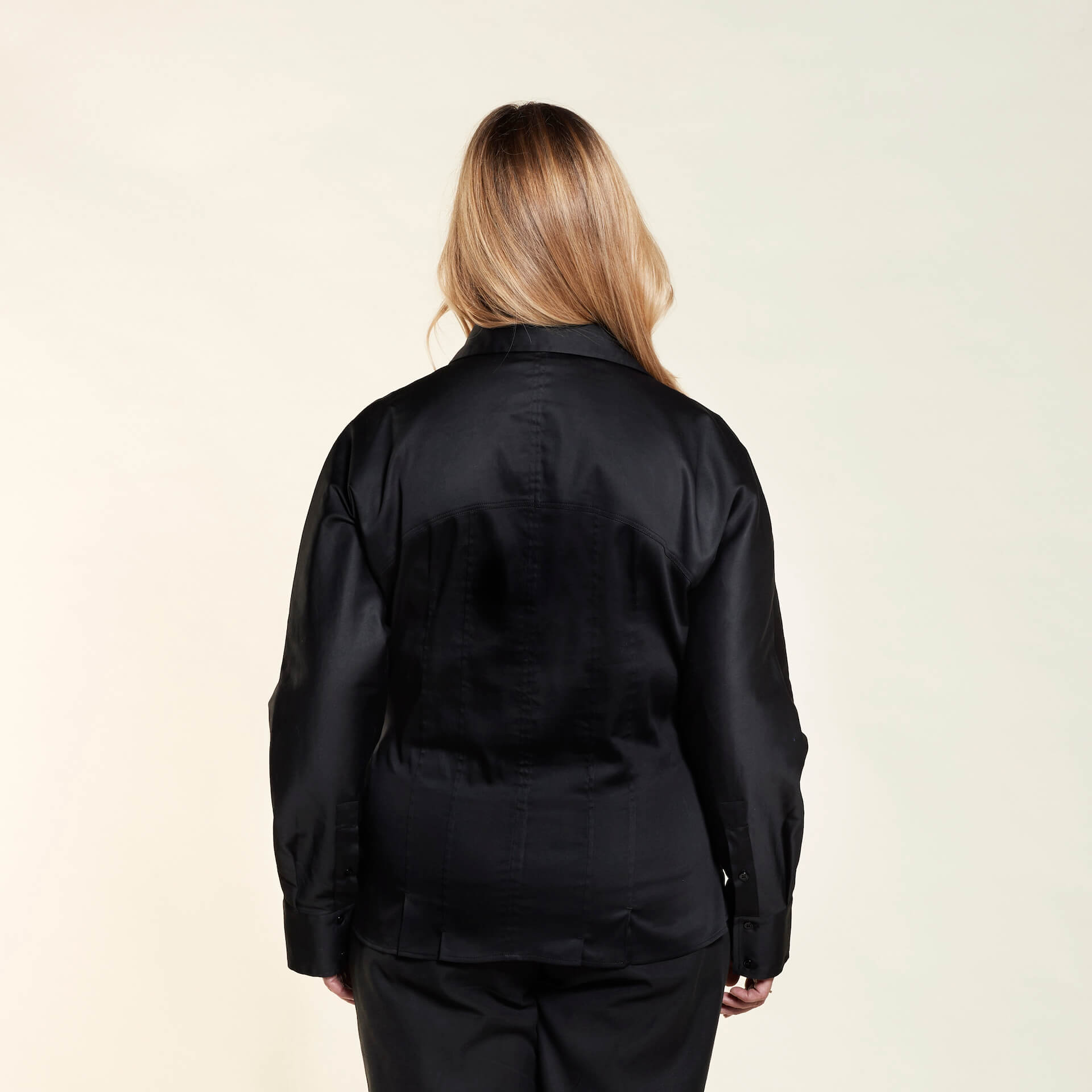 Rear view of a woman with blonde hair wearing a black button-up shirt and black pants. She is standing against a light background, facing away from the camera.