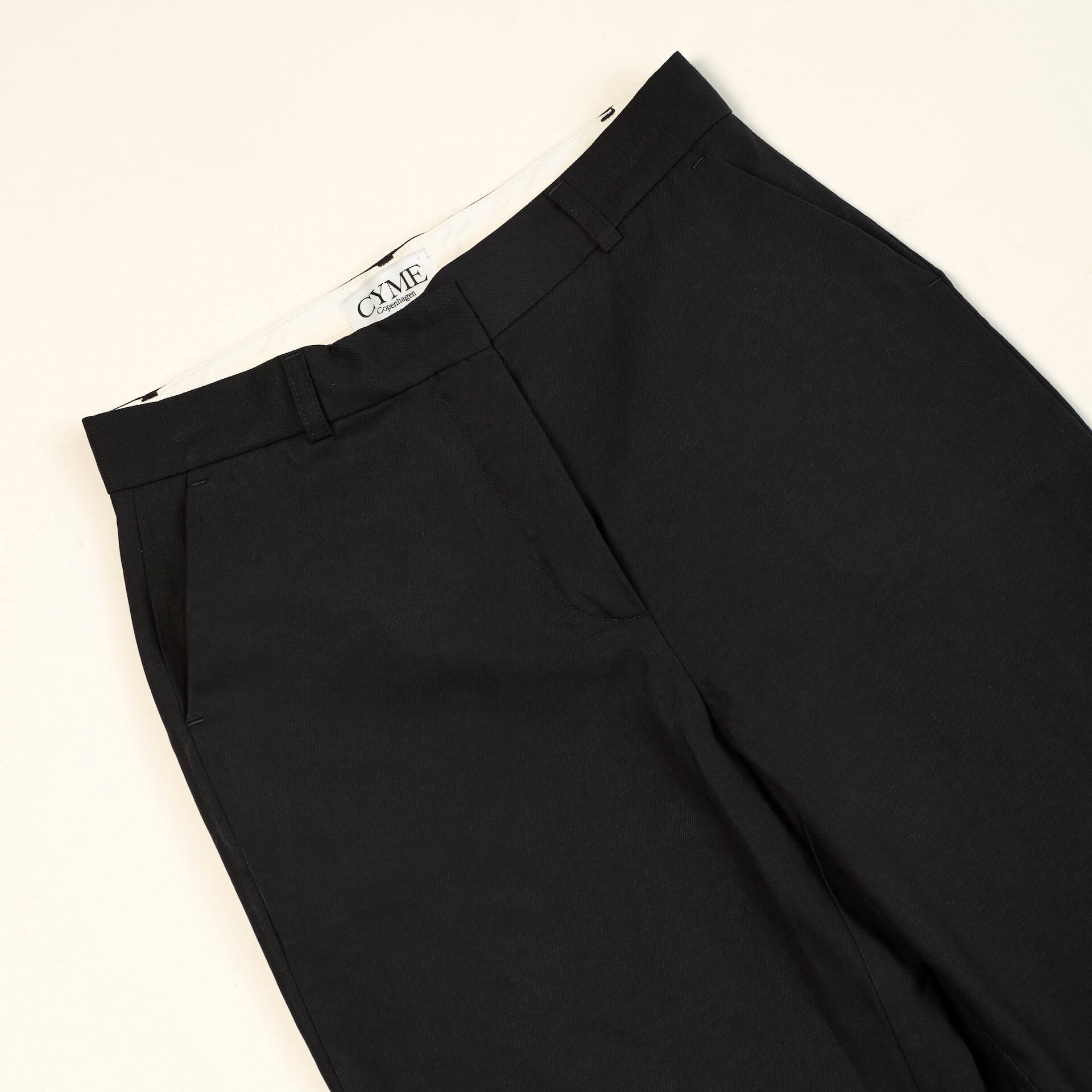 Close-up of a pair of black trousers from CYME Copenhagen. The trousers feature a sleek, minimalistic design with a high waist and side pockets. The CYME Copenhagen label is prominently displayed on the inner waistband.