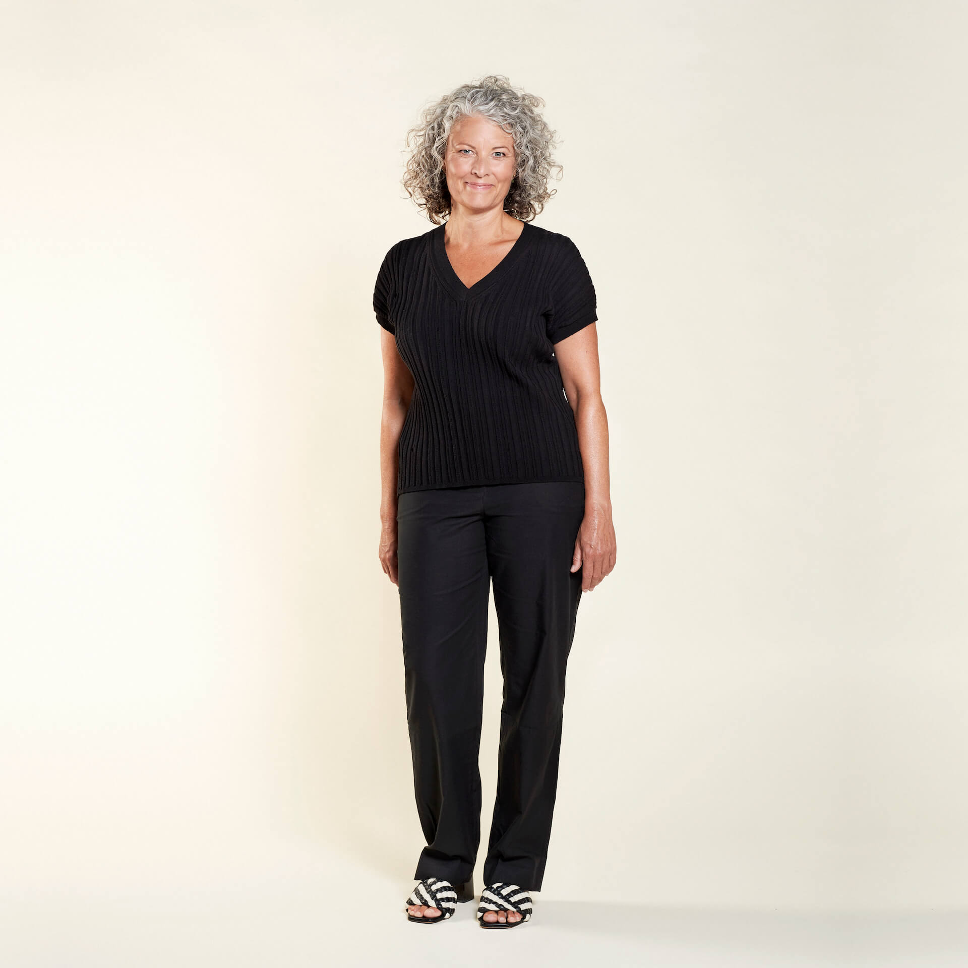 A woman with curly gray hair models a classic outfit from CYME Copenhagen. She is wearing a black V-neck short-sleeve sweater paired with black trousers. The look is completed with black and white striped sandals, emphasizing the brand's focus on timeless, high-quality fashion for all ages.