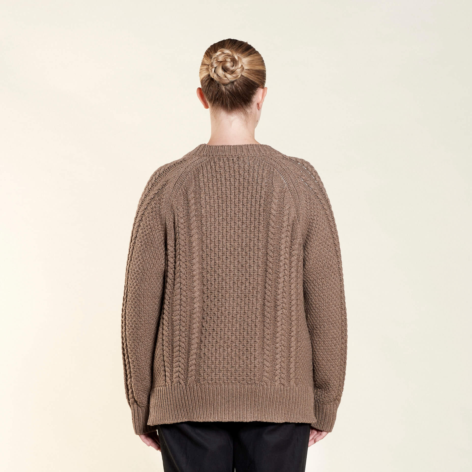 Rear view of a woman with her hair styled in a braided bun, modeling a chunky brown cable-knit cardigan from CYME Copenhagen. The image highlights the intricate knit pattern of the cardigan, paired with black trousers, showcasing the brand's focus on comfort and timeless design.