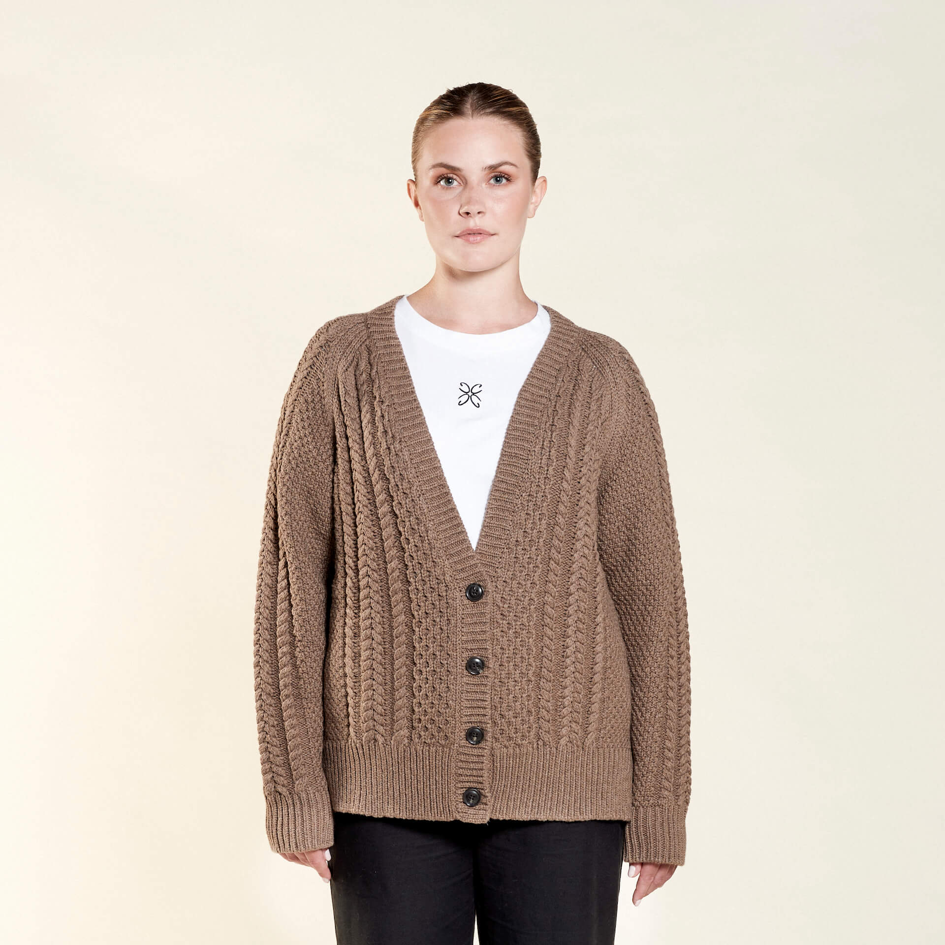 A woman models a cozy outfit from CYME Copenhagen. She is wearing a chunky brown cable-knit cardigan over a white top with an embroidered logo, paired with black trousers. The look emphasizes comfort and the brand's commitment to timeless, high-quality fashion.