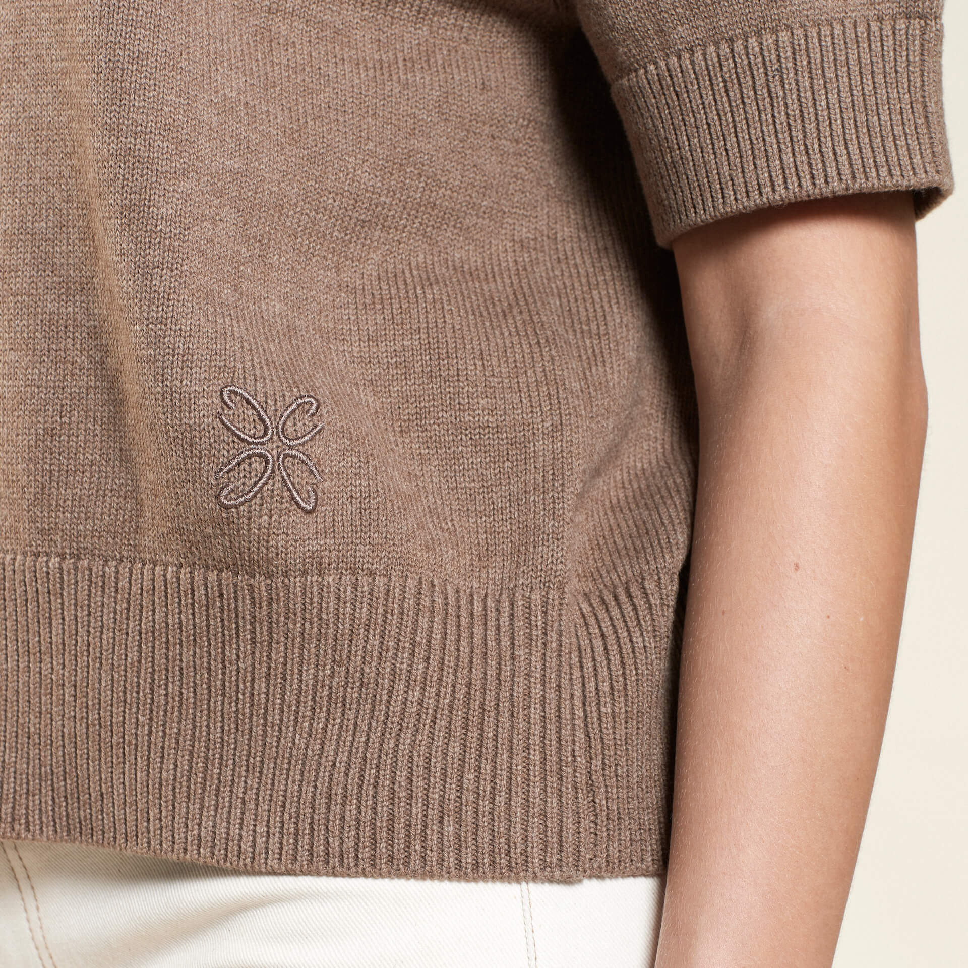 Close-up of a brown knit sweater from CYME Copenhagen, featuring a subtle embroidered logo on the lower side, paired with light-colored trousers. The image highlights the fine details and craftsmanship of the garment.