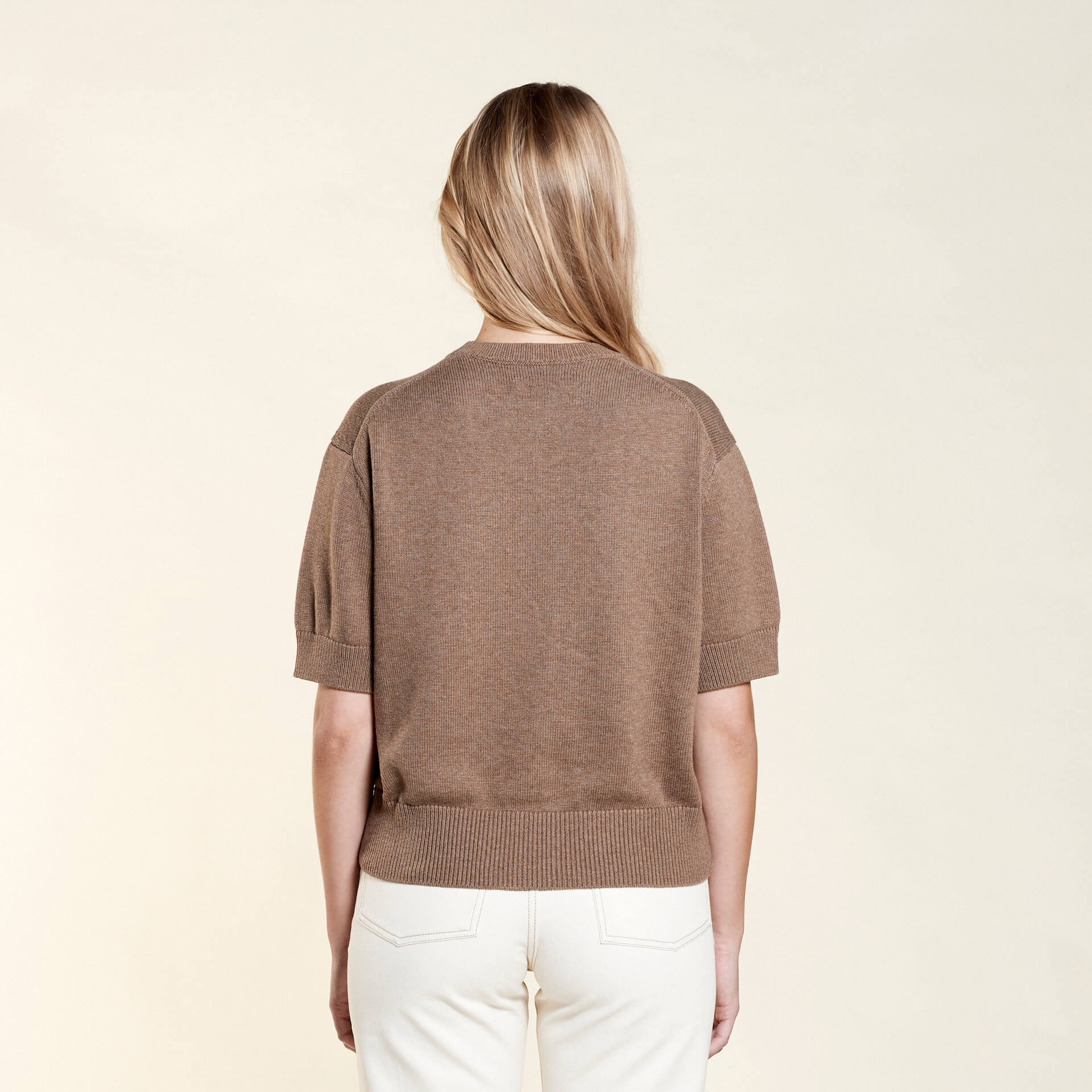 Rear view of a woman with long blonde hair modeling a brown short-sleeve sweater paired with off-white jeans from CYME Copenhagen. The image showcases the sweater's relaxed fit and the clean lines of the outfit, emphasizing the brand's focus on timeless, versatile fashion.