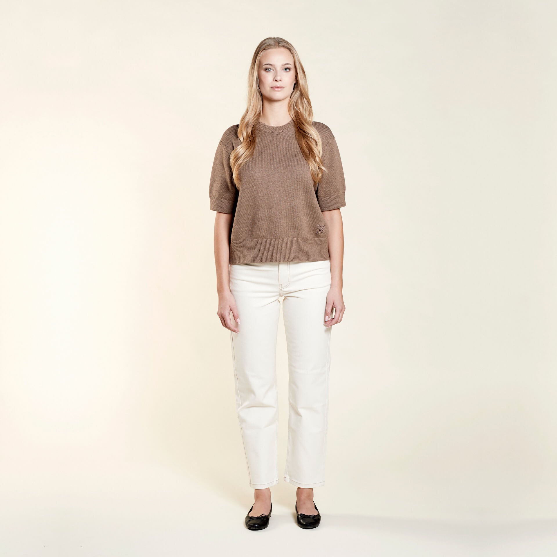 A woman with long blonde hair models a casual outfit from CYME Copenhagen. She is wearing a brown short-sleeve sweater paired with off-white jeans. The look is completed with black ballet flats, emphasizing the brand's focus on timeless, high-quality fashion.