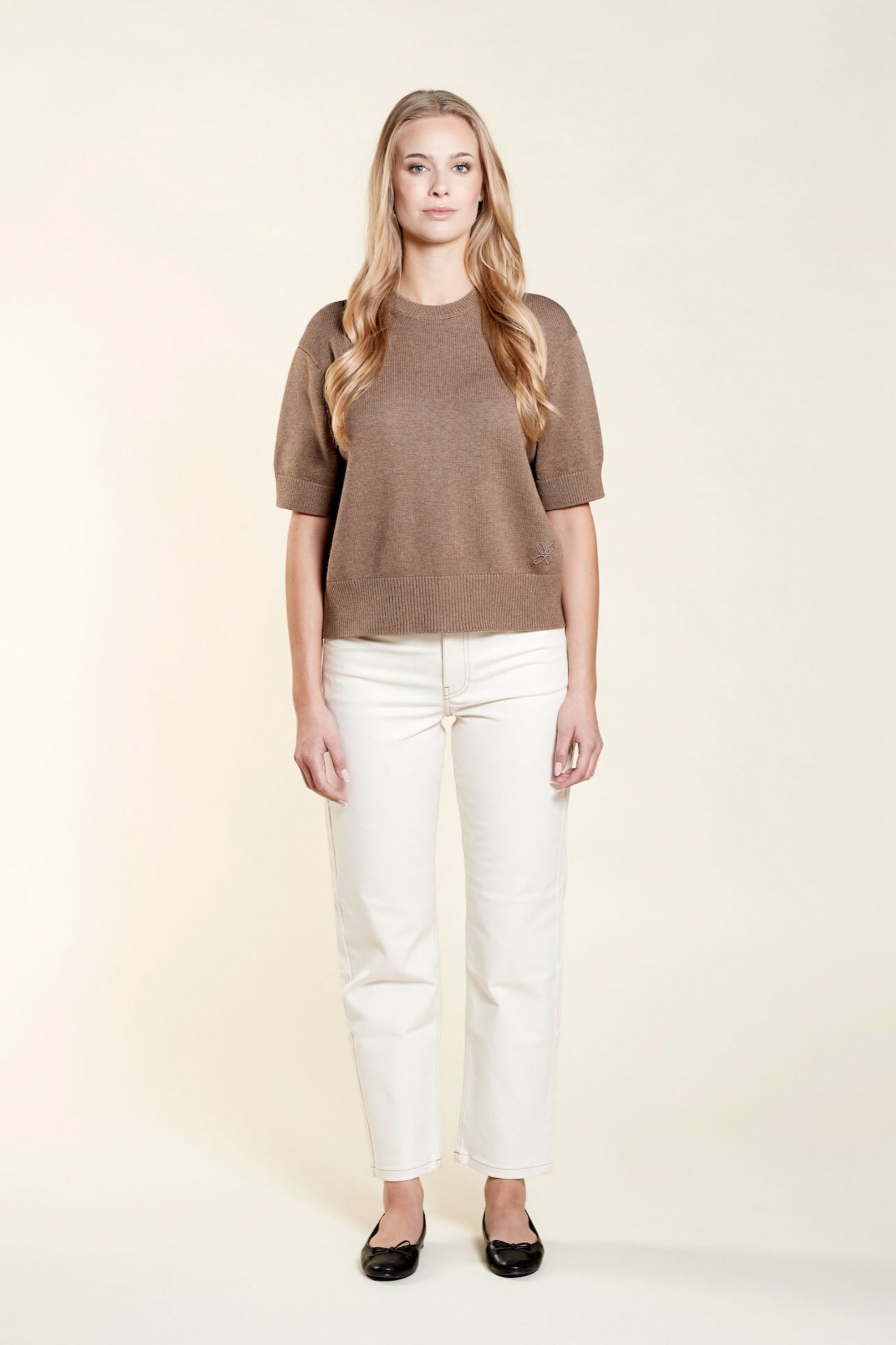 A woman with long blonde hair models a casual outfit from CYME Copenhagen. She is wearing a brown short-sleeve sweater paired with off-white jeans. The look is completed with black ballet flats, emphasizing the brand's focus on timeless, high-quality fashion.