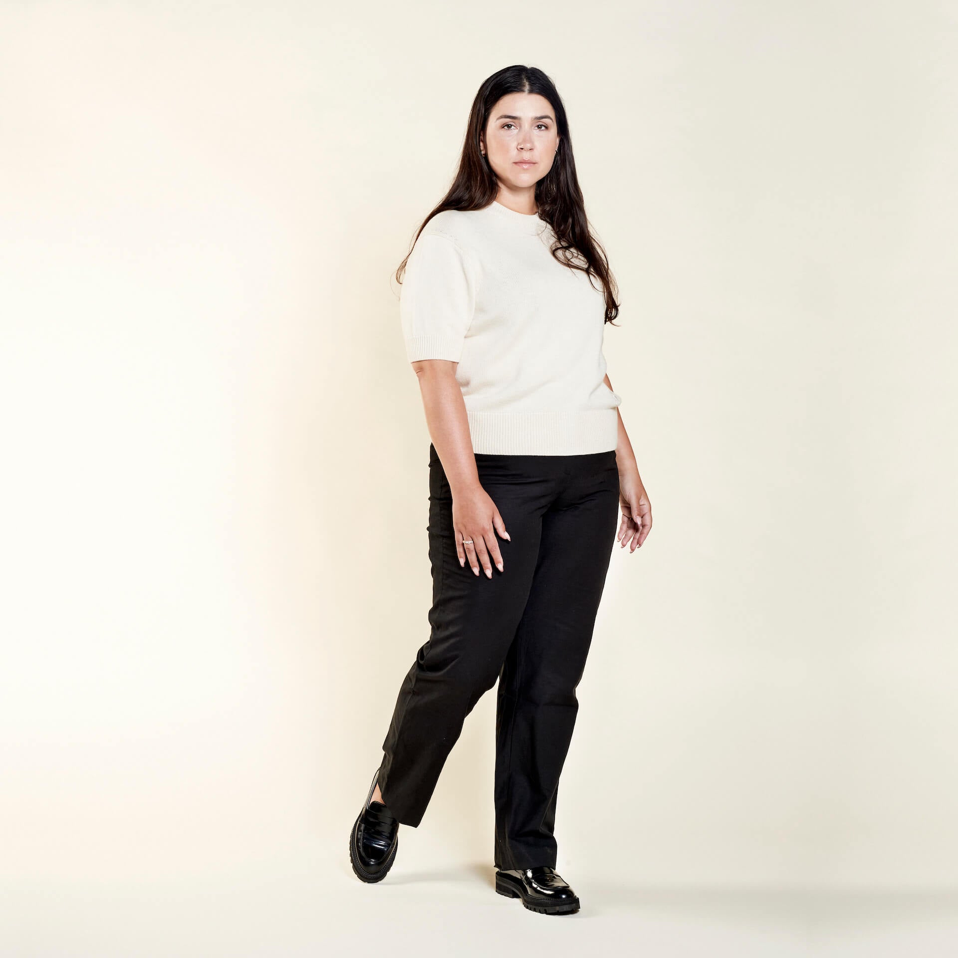 A woman with long dark hair models a casual outfit from CYME Copenhagen. She is wearing a cream-colored short-sleeve sweater paired with black trousers. The look is completed with black loafers, emphasizing the brand's focus on timeless, high-quality fashion.