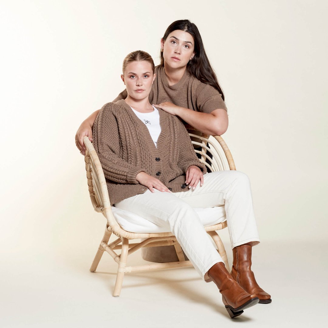 Two women model a cozy, casual look from CYME Copenhagen. One woman is seated in a wicker chair, wearing a chunky brown cardigan over a white top paired with white jeans and brown boots. The other woman stands behind her, wearing a brown short-sleeve sweater, showcasing the brand's focus on high-quality, timeless fashion.