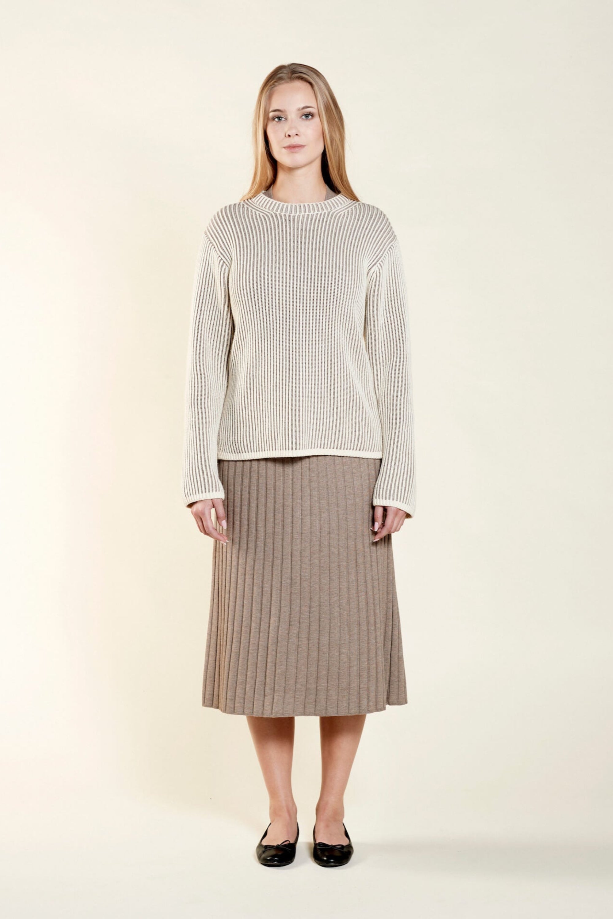 Portrait of a woman with long blonde hair wearing a cream-colored ribbed sweater and a beige pleated skirt. She is standing against a light background, looking straight at the camera with a neutral expression.