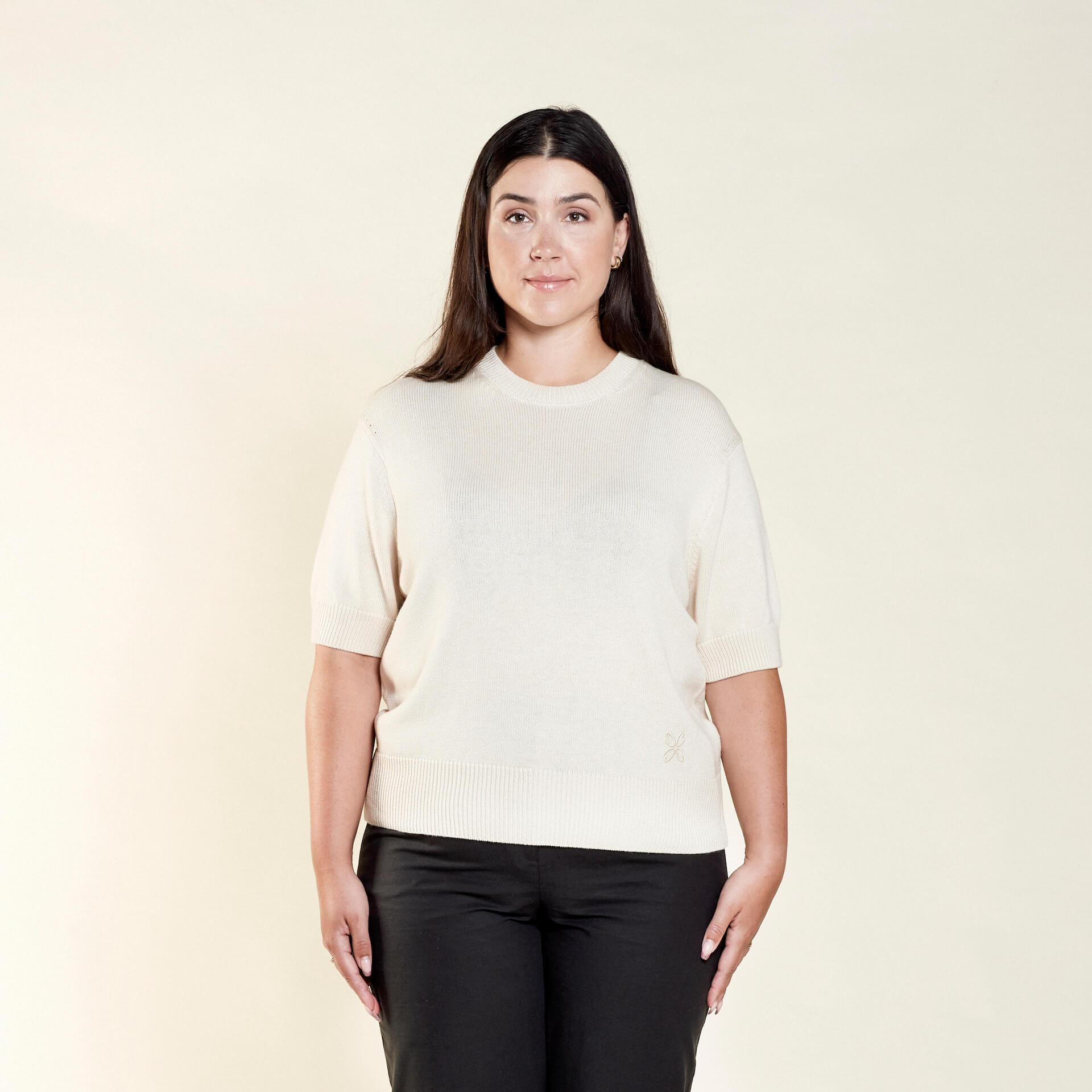 A woman with long dark hair models a cream-colored short-sleeve sweater paired with black trousers from CYME Copenhagen. The outfit emphasizes the brand's commitment to timeless, versatile, and comfortable fashion.