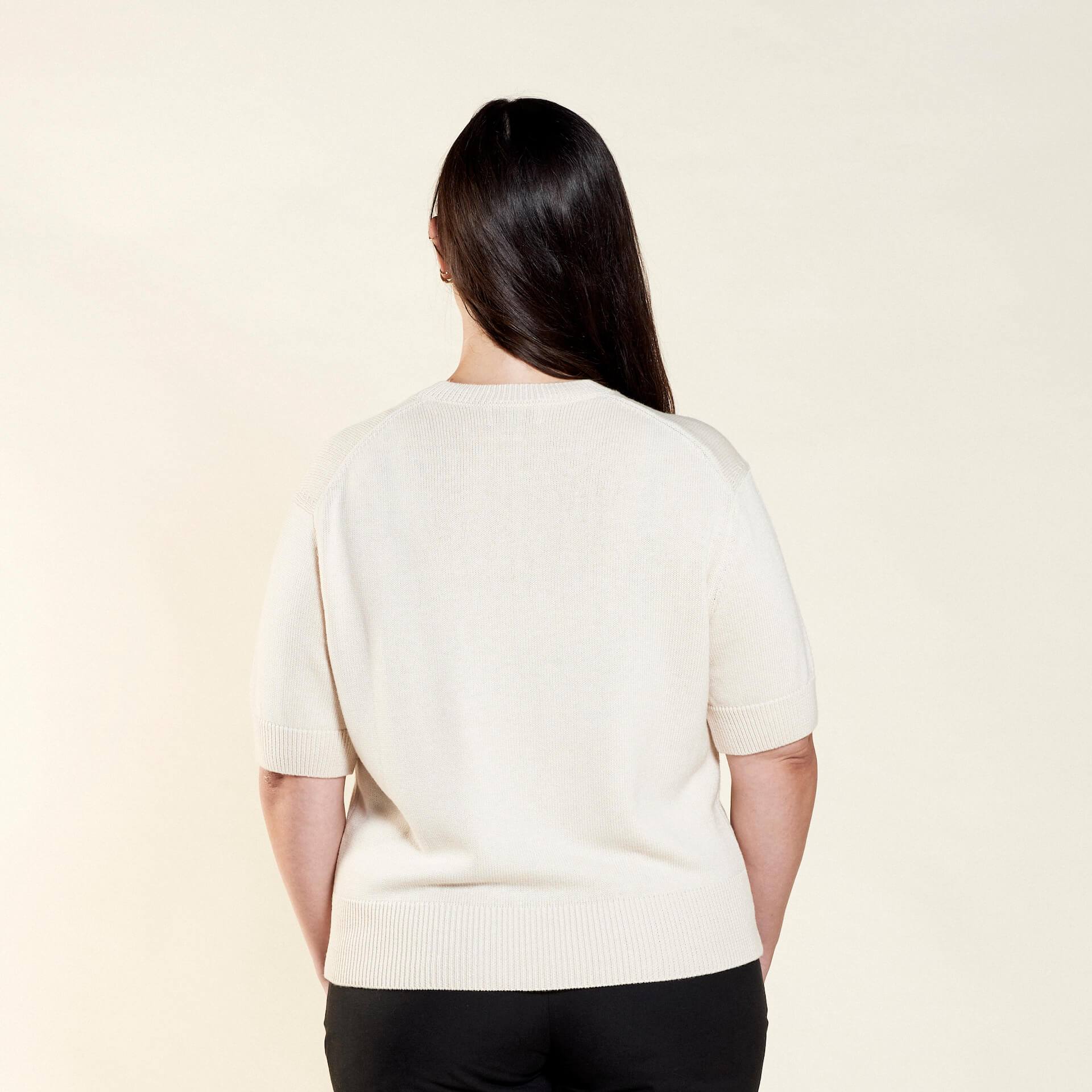 Rear view of a woman with long dark hair modeling a cream-colored short-sleeve sweater from CYME Copenhagen. The image showcases the sweater's relaxed fit and clean design, paired with black trousers, highlighting the brand's focus on timeless, versatile fashion.