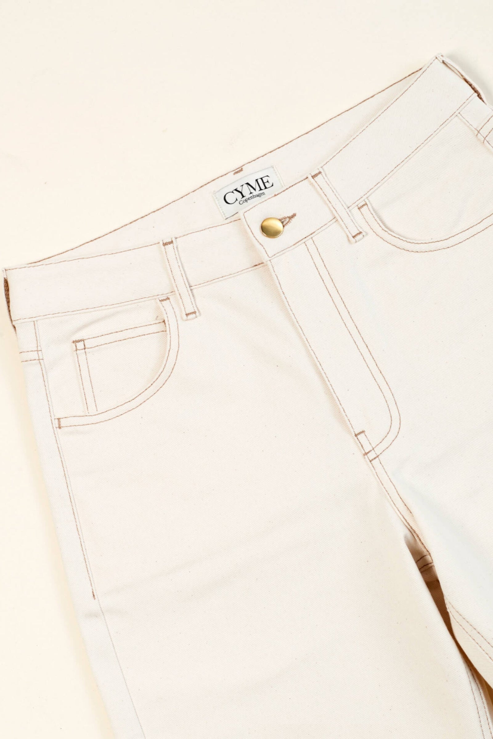 Close-up of a pair of beige high-waisted jeans from CYME Copenhagen. The jeans feature a classic five-pocket design, with visible stitching and a brass button closure. The CYME Copenhagen label is prominently displayed on the inner waistband.