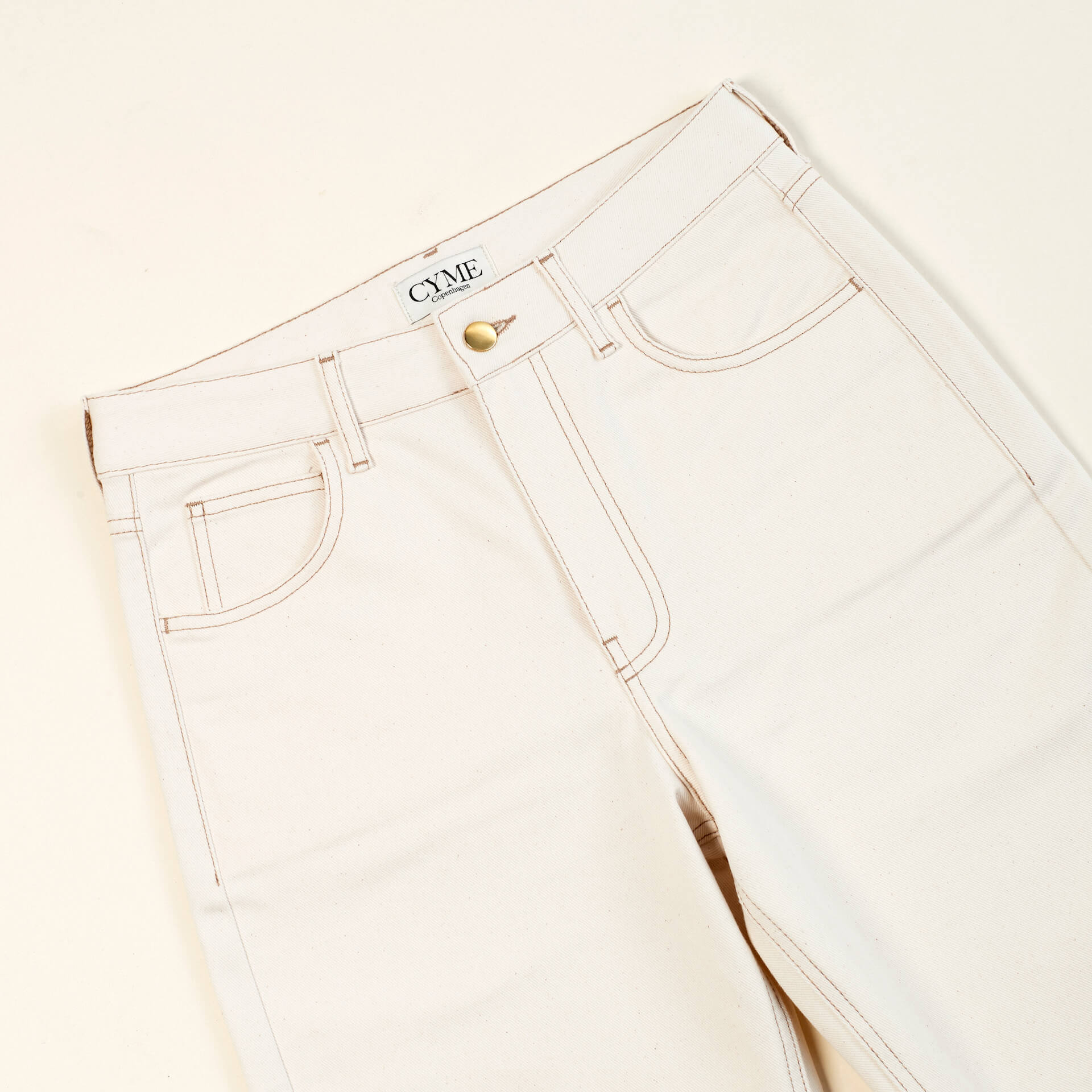 Close-up of a pair of beige high-waisted jeans from CYME Copenhagen. The jeans feature a classic five-pocket design, with visible stitching and a brass button closure. The CYME Copenhagen label is prominently displayed on the inner waistband.