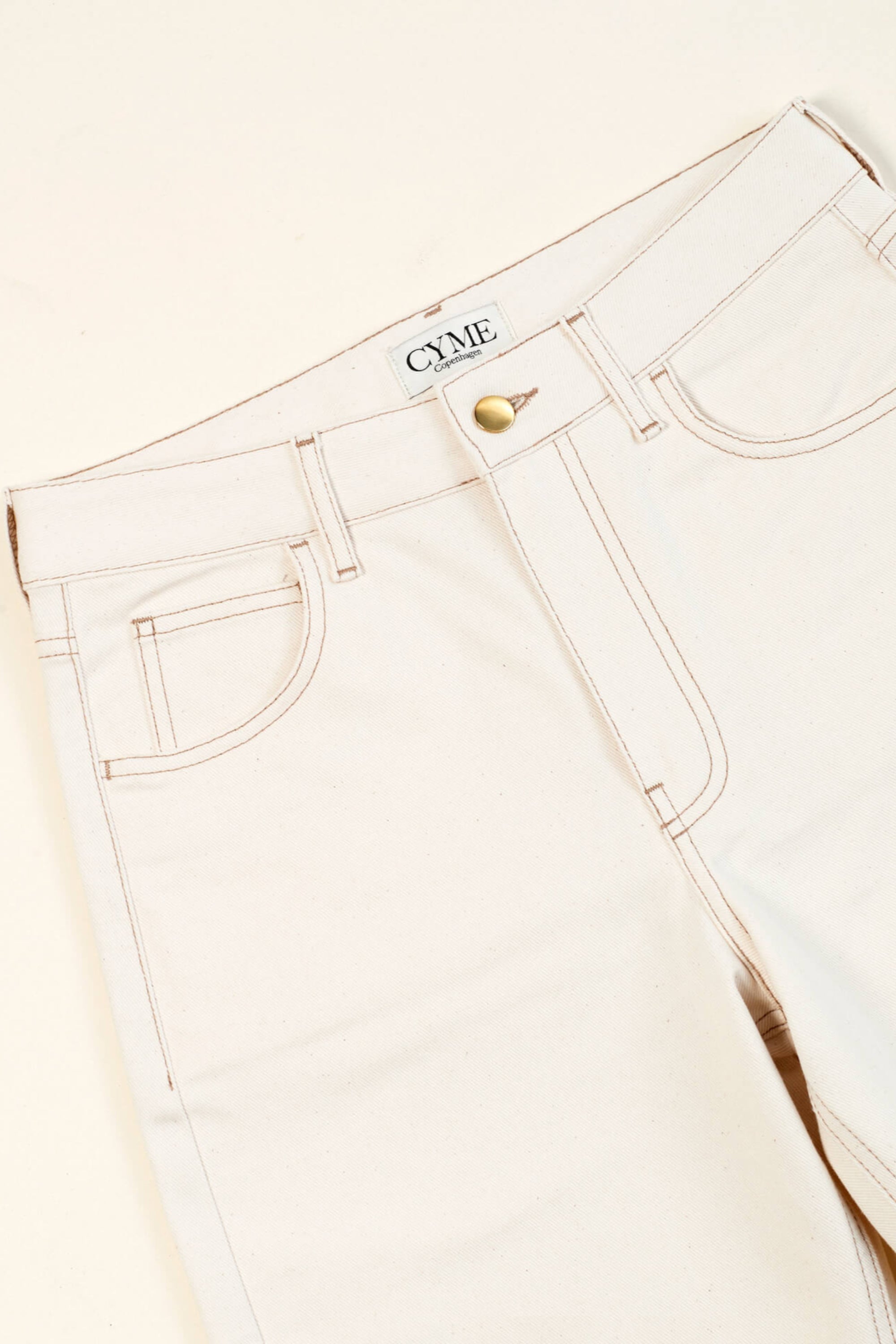 Close-up of a pair of beige high-waisted jeans from CYME Copenhagen. The jeans feature a classic five-pocket design, with visible stitching and a brass button closure. The CYME Copenhagen label is prominently displayed on the inner waistband.