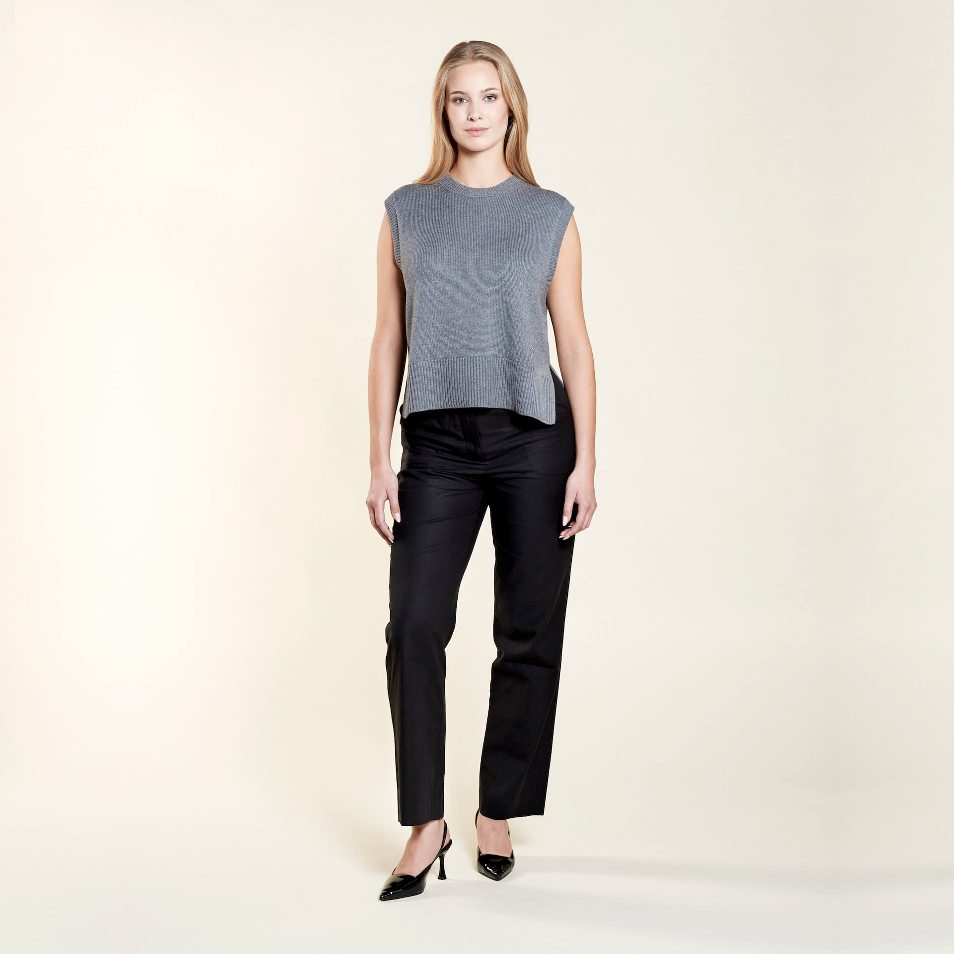 A woman with long blonde hair models a chic outfit from CYME Copenhagen. She is wearing a sleeveless gray knit top paired with black trousers and black heels, showcasing a minimalist and sophisticated look that emphasizes the brand's commitment to timeless fashion.