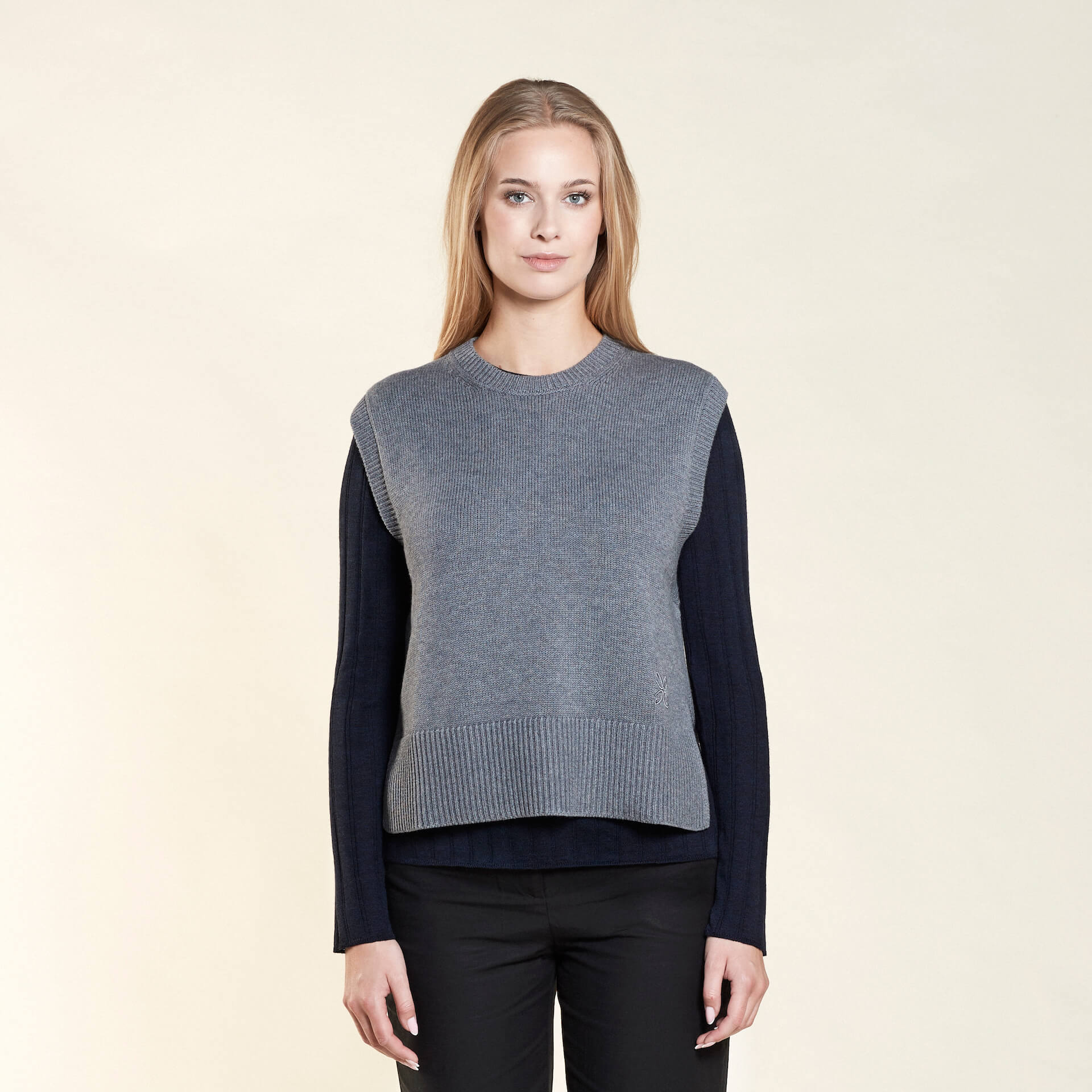 A woman with long blonde hair models a gray sleeveless knit vest layered over a navy blue long-sleeve top, paired with black trousers from CYME Copenhagen. The outfit highlights the brand's focus on timeless layering and versatile fashion.