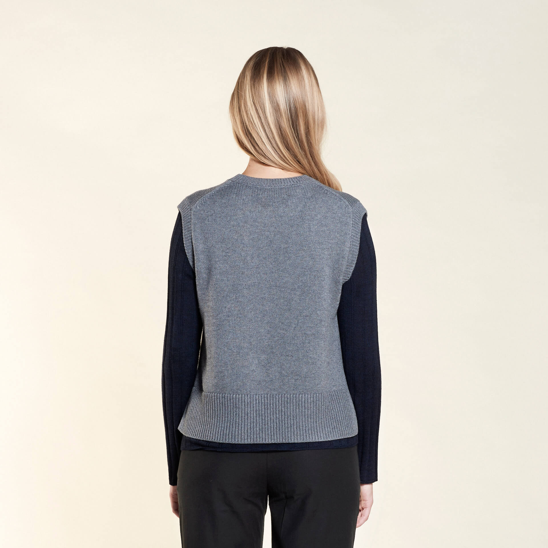 Rear view of a woman with long blonde hair modeling a gray sleeveless knit vest layered over a navy blue long-sleeve top, paired with black trousers from CYME Copenhagen. The image highlights the vest's relaxed fit and the layered look, emphasizing the brand's focus on timeless, versatile fashion.