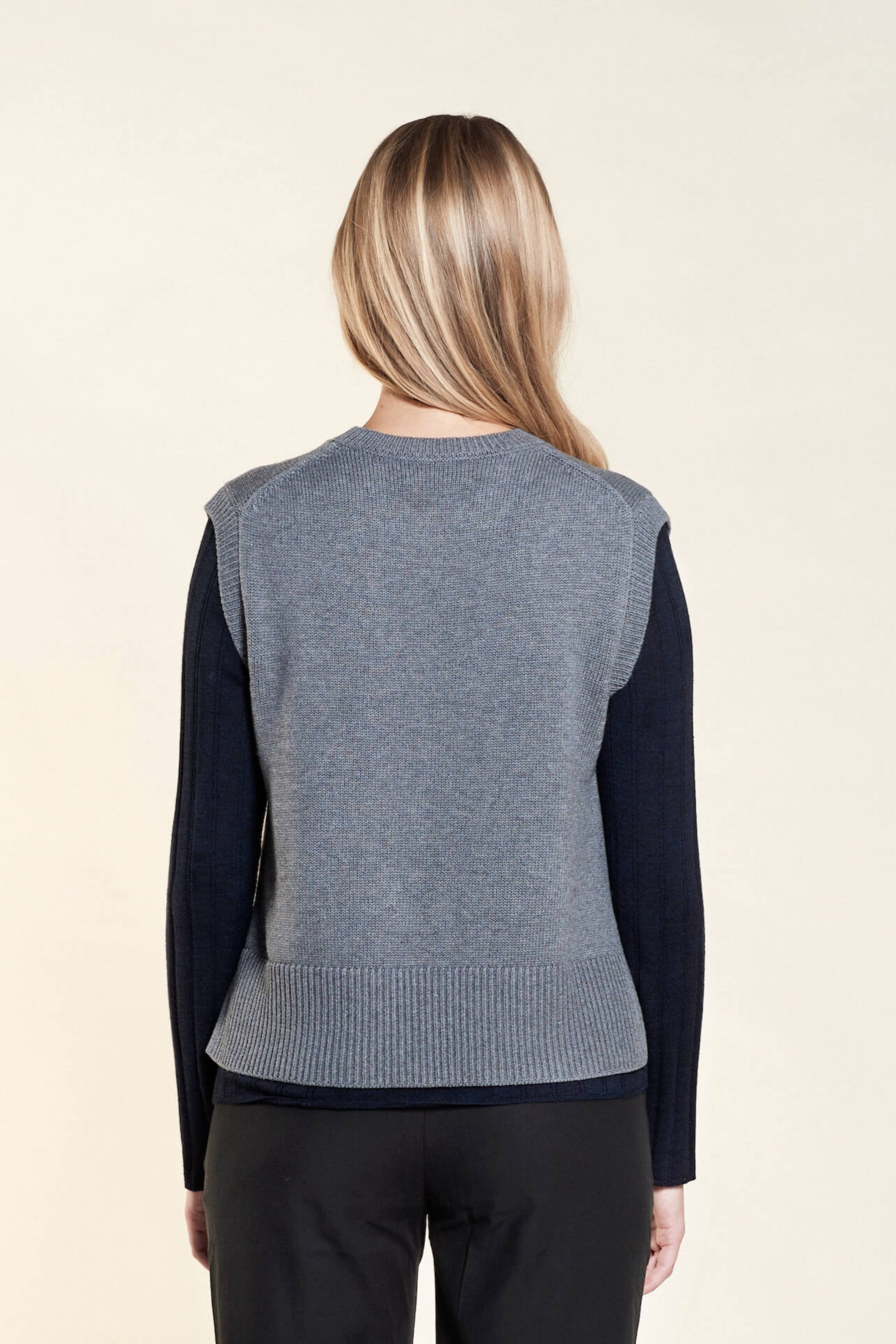 Rear view of a woman with long blonde hair modeling a gray sleeveless knit vest layered over a navy blue long-sleeve top, paired with black trousers from CYME Copenhagen. The image highlights the vest's relaxed fit and the layered look, emphasizing the brand's focus on timeless, versatile fashion.