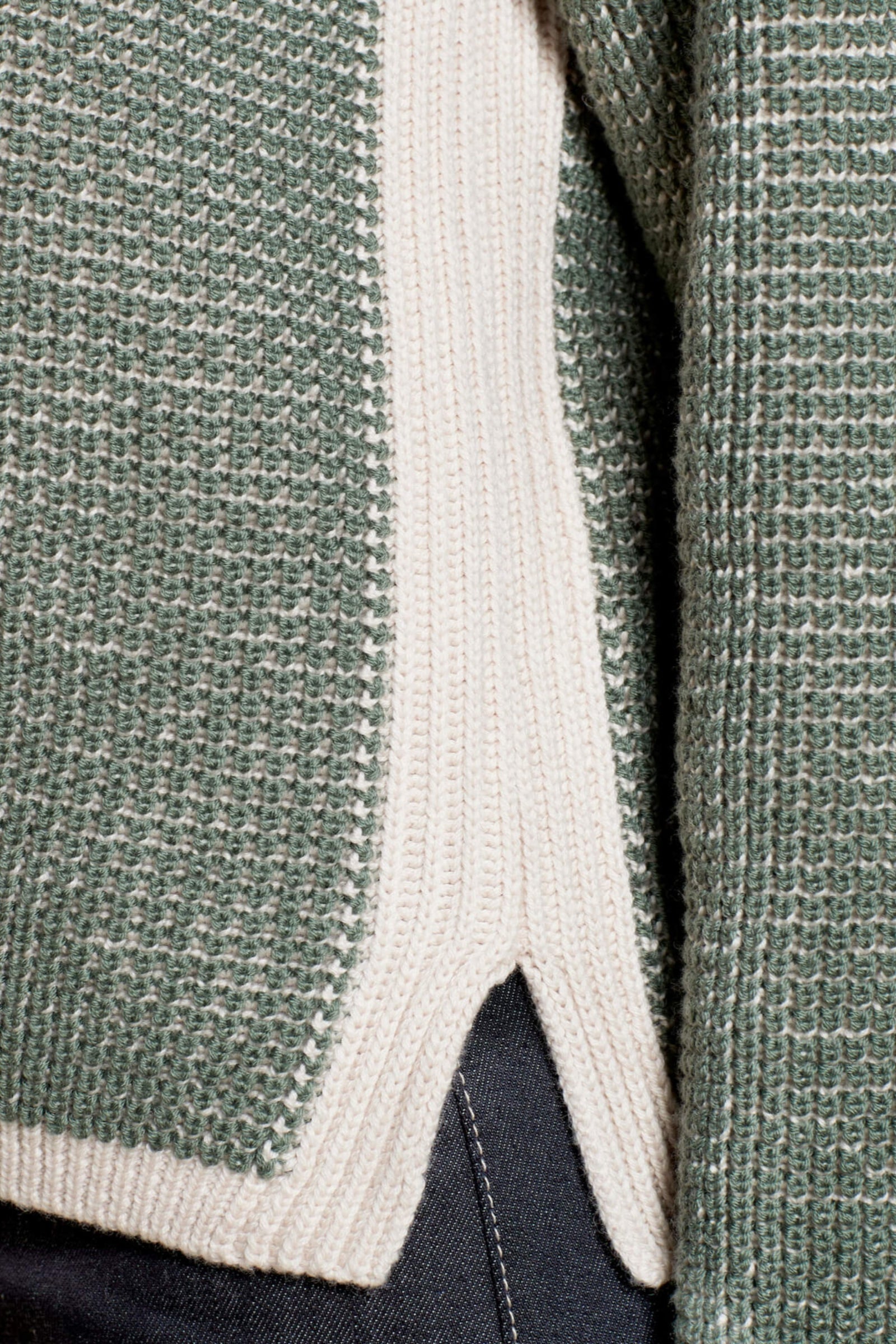 Close-up of a green knit sweater with cream accents at the hem, highlighting the texture and intricate weave of the fabric. The focus is on the side slit and layered design of the garment, showing part of the underlying cream knit panel.