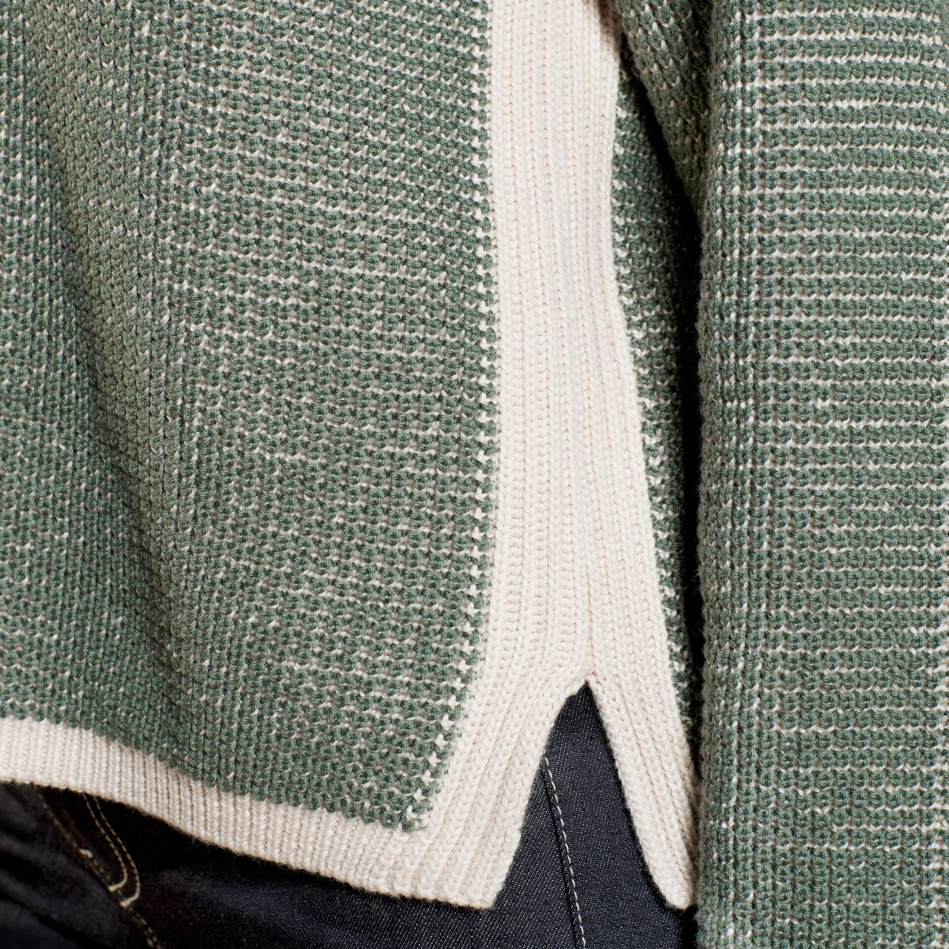 Close-up of a green knit sweater with cream accents at the hem, highlighting the texture and intricate weave of the fabric. The focus is on the side slit and layered design of the garment, showing part of the underlying cream knit panel.